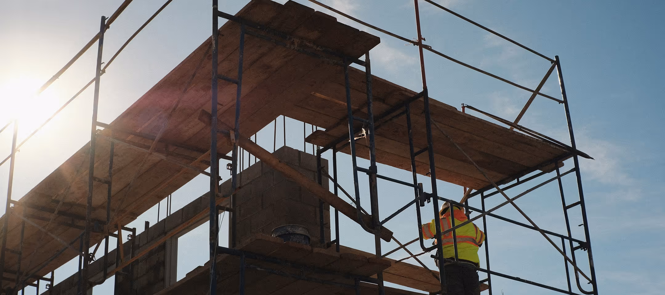 Construction worker in a yellow safety jacket working on metal scaffolding under a clear blue sky with the sun shining.