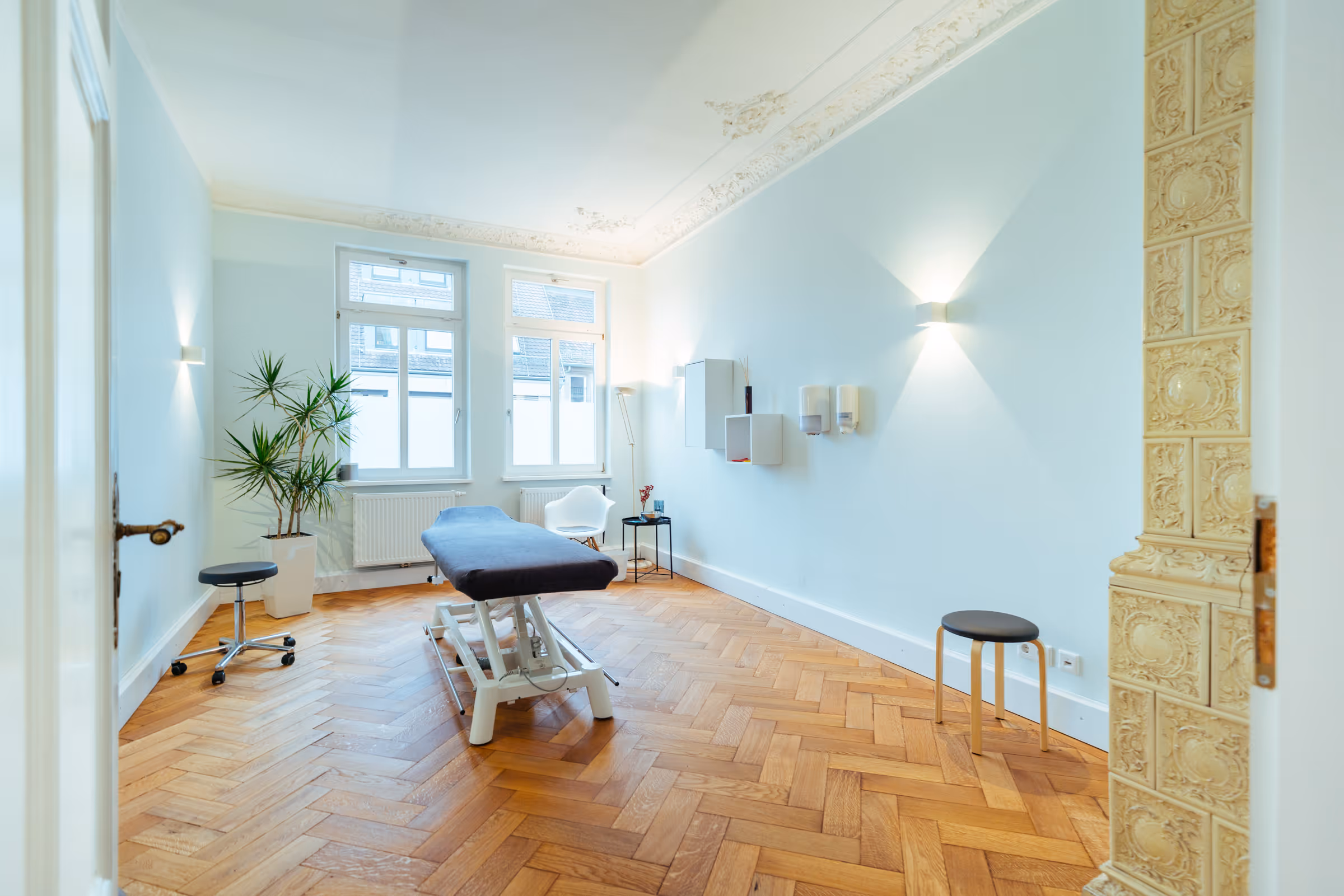 Bright treatment room with wooden herringbone floor, black cushioned therapy table, white chair, potted plant, and large windows.