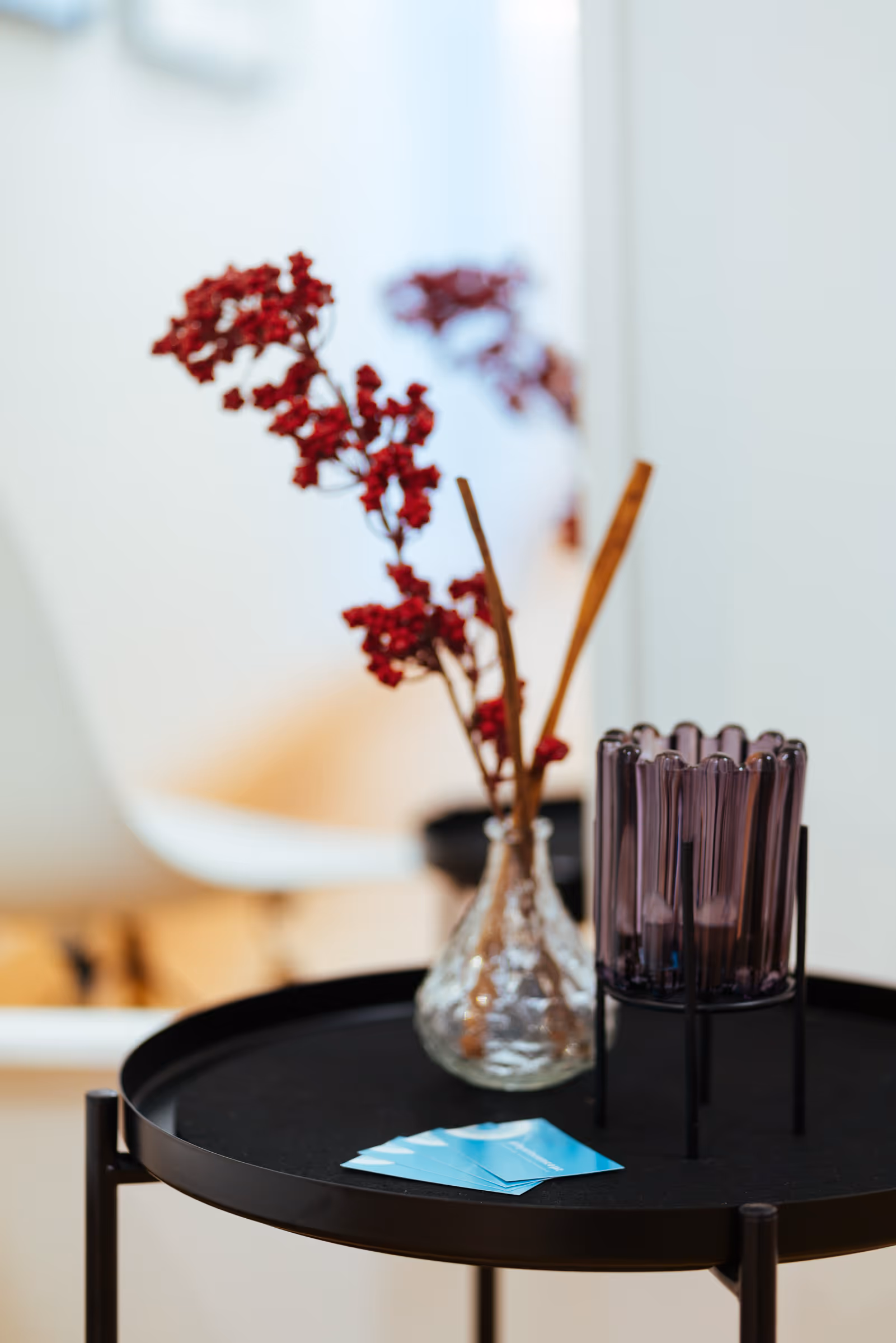 Black round side table with a clear glass vase holding red dried flowers and a purple glass candle holder on it.