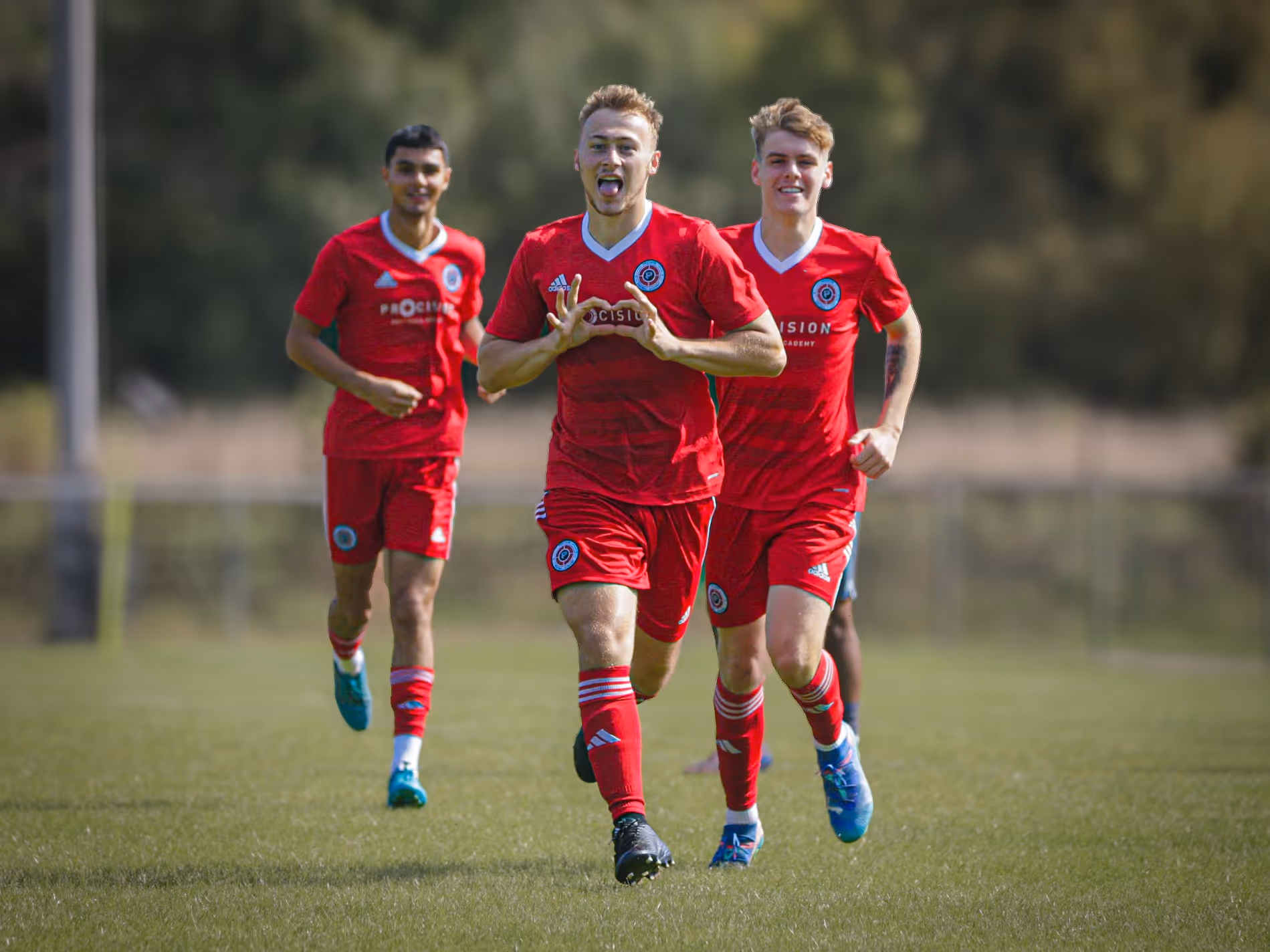 Three soccer players in red uniforms running on a grass field, one making a hand heart gesture.