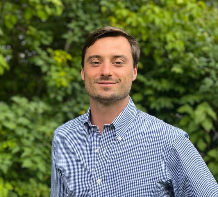 Man with short brown hair wearing a blue and white checkered shirt standing outdoors with green leafy background.