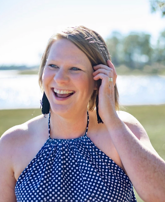 Smiling woman with short hair wearing a blue polka dot halter top and black tassel earrings outdoors near water.