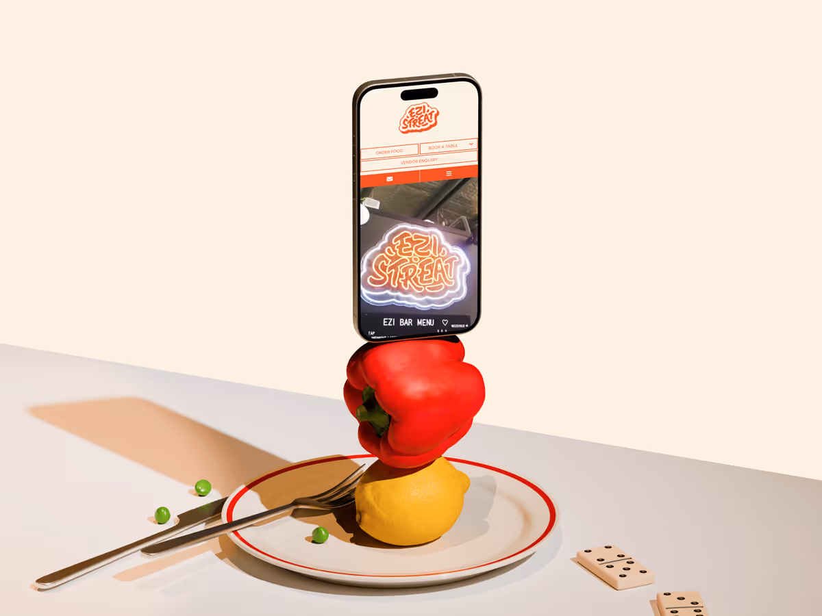 Smartphone displaying Ezi Streat website balanced vertically on a red bell pepper and a lemon on a white plate with scattered green peas, fork, and dominoes nearby.