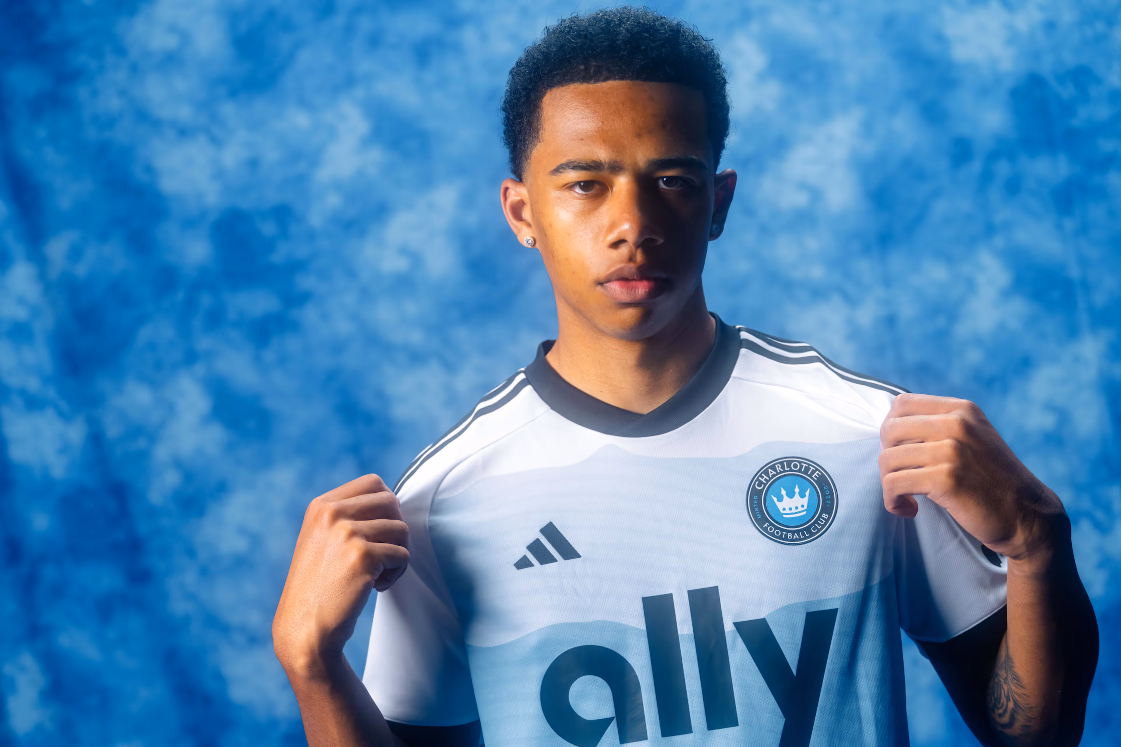 Young male athlete holding the front of a Charlotte Football Club jersey with a blue and white background.