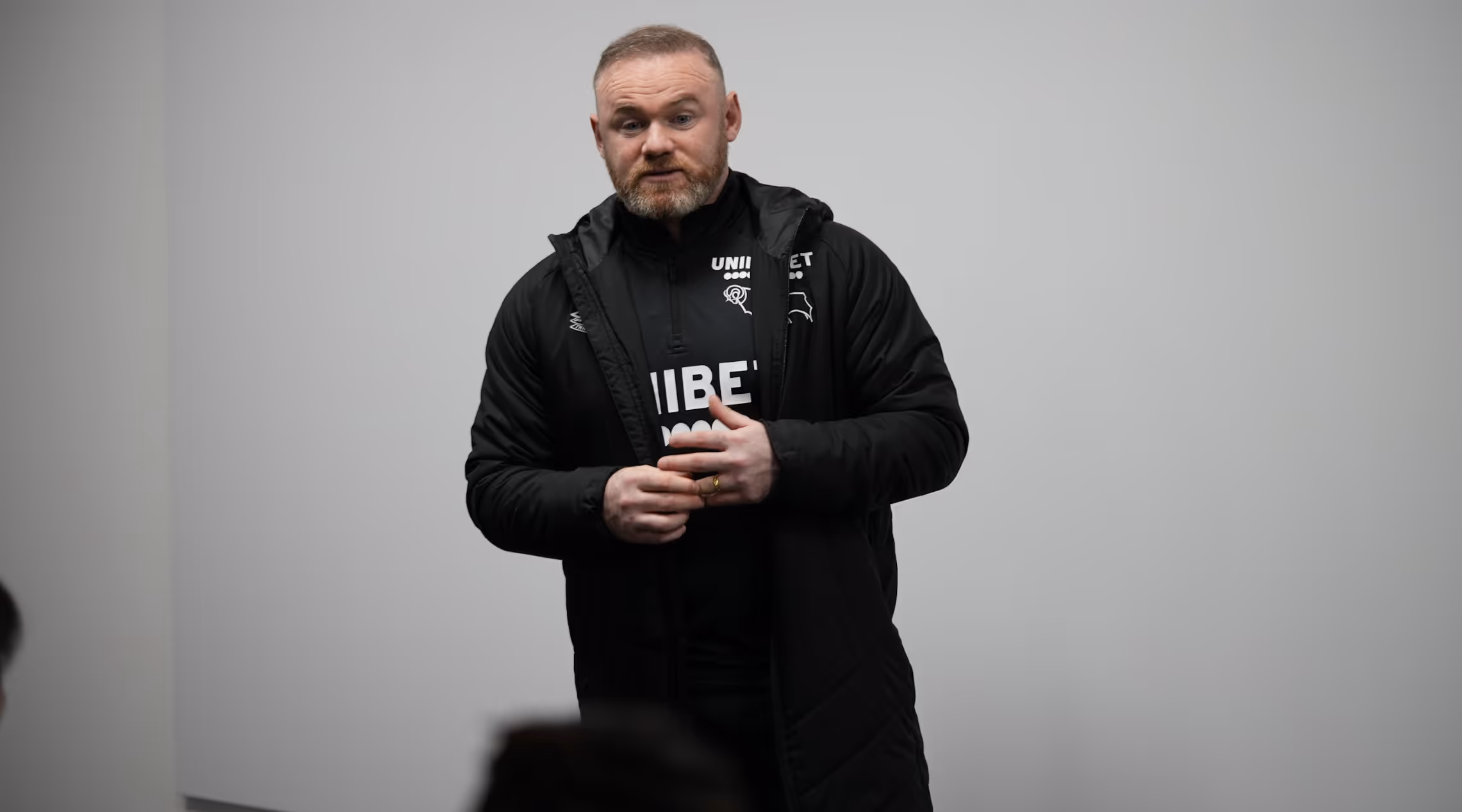 Man with a beard wearing a black jacket and black sportswear, standing against a plain light gray background.