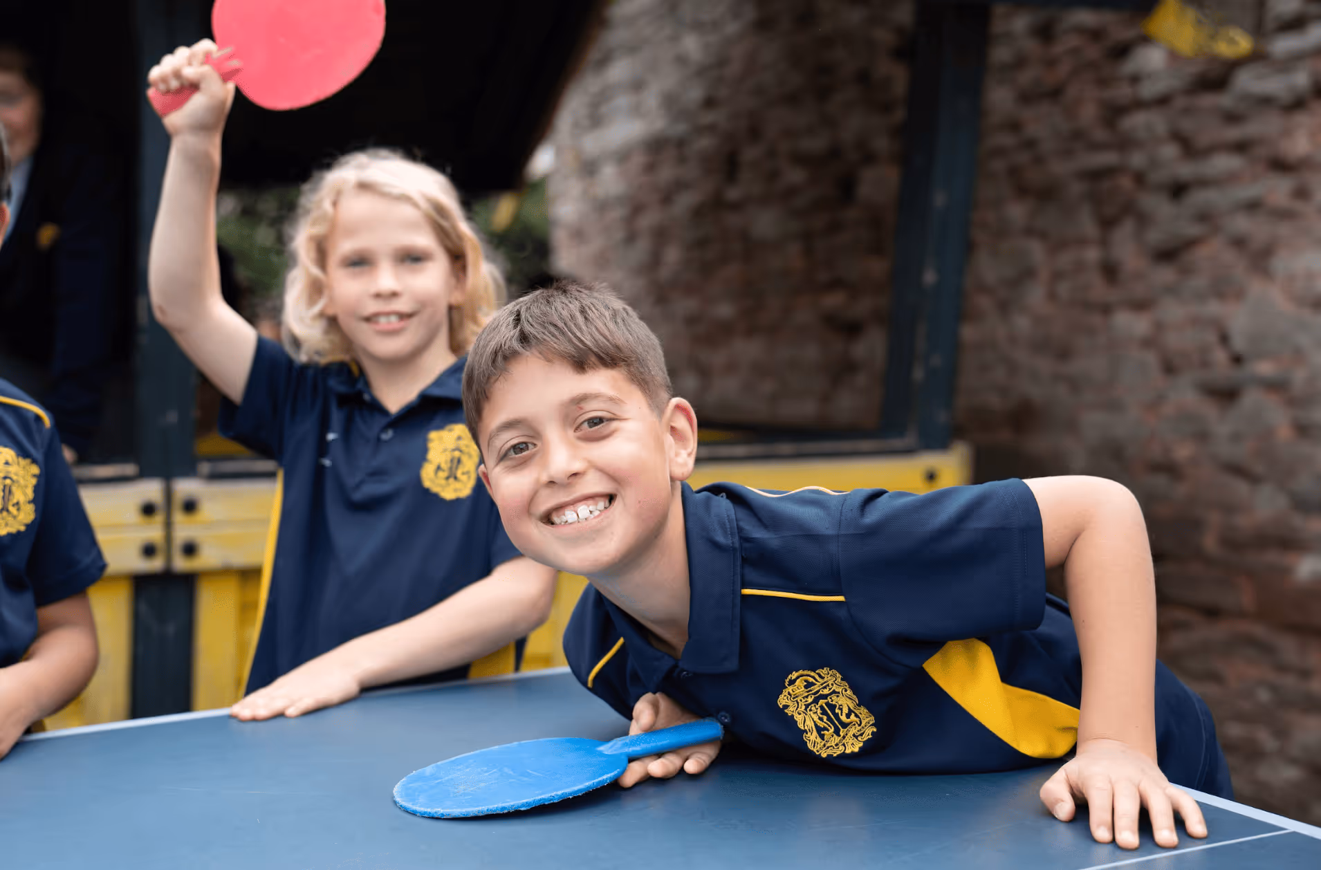 Two children in navy and yellow school uniforms playing table tennis outdoors, one holding a blue paddle and smiling at the camera, the other raising a red paddle.