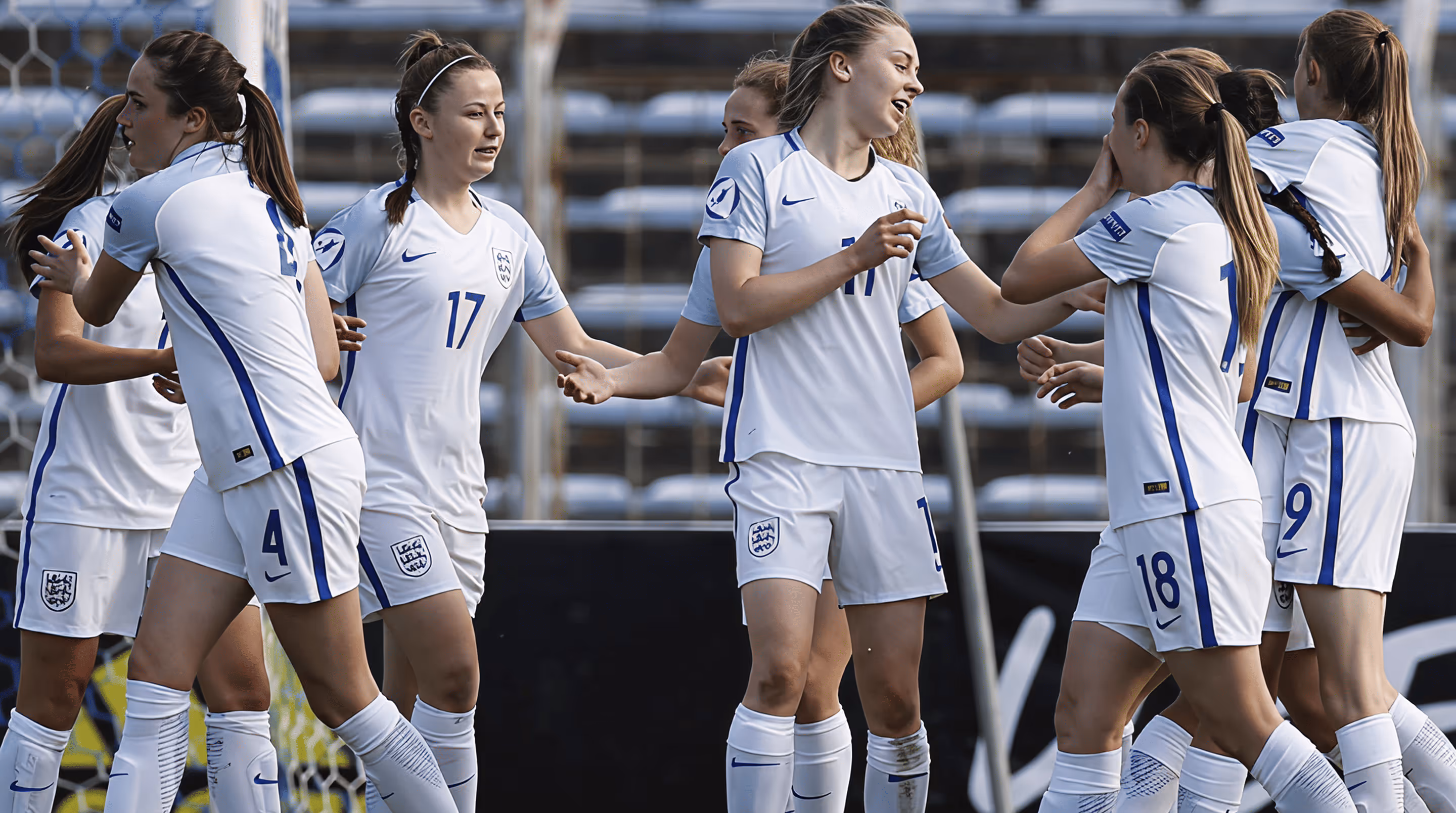 Women soccer players in white and blue uniforms celebrating on the field near the goalpost.