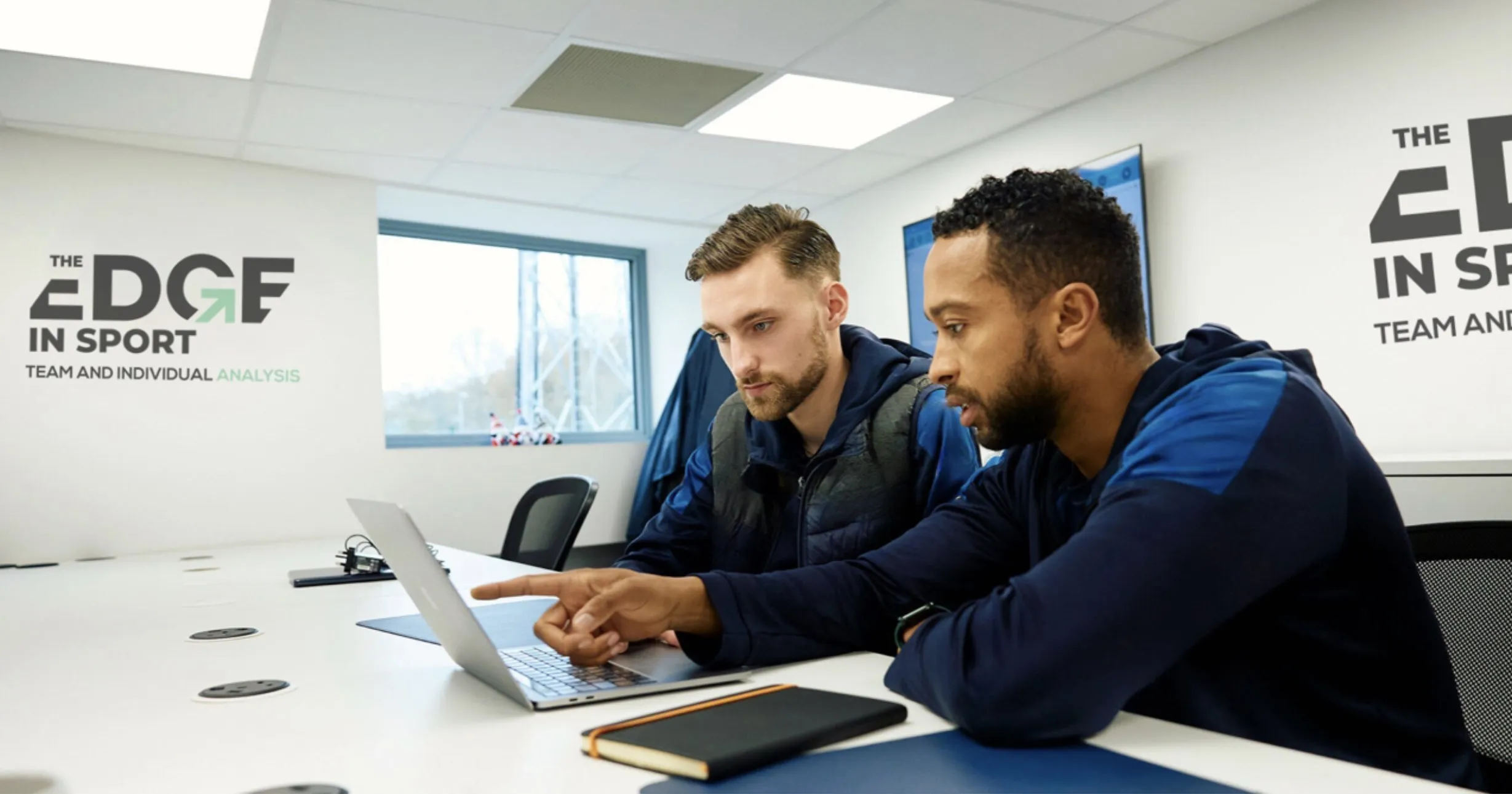 Two men seated at a conference table looking at a laptop, with 'The Edge in Sport Team and Individual Analysis' text on the wall behind them.