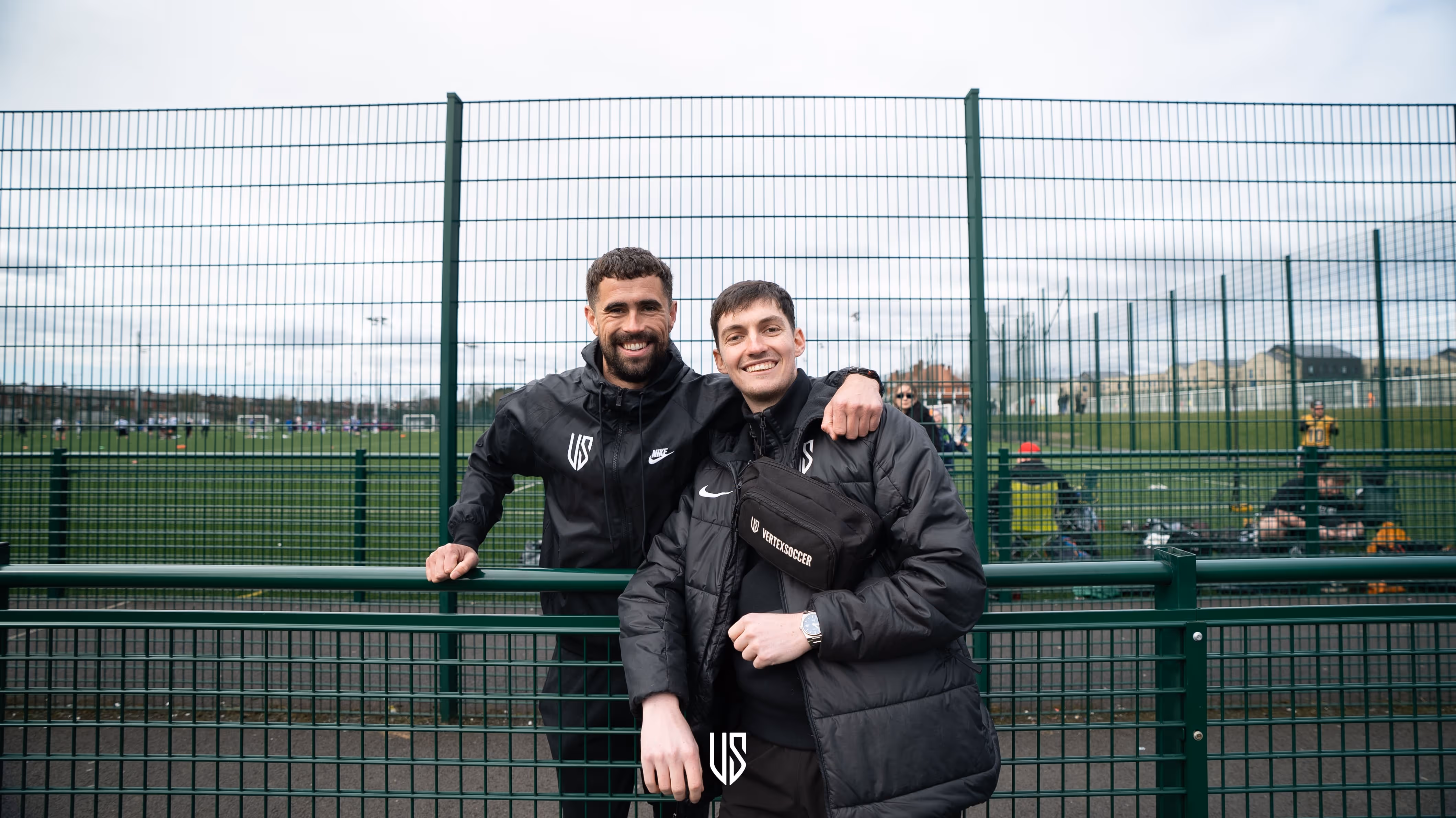 Two men in black jackets smiling and posing in front of a green metal fence at a soccer field.