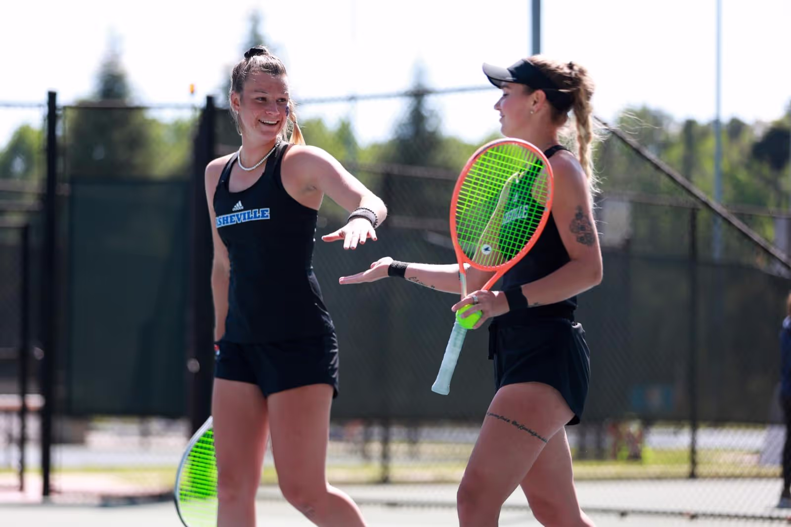 Two female tennis players in black outfits sharing a light moment on an outdoor court during daytime.