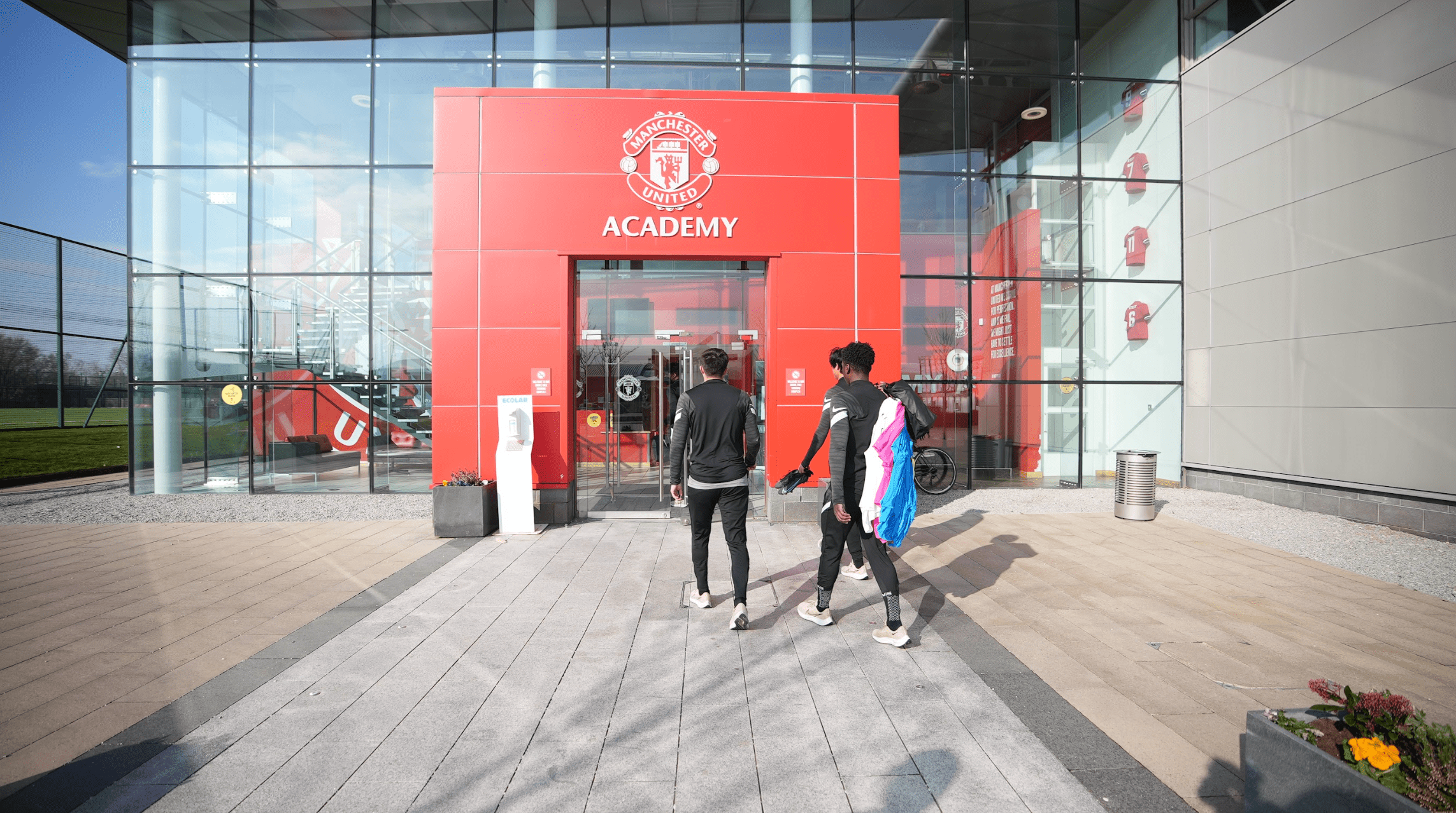 Three young men in black sportswear walking toward the red entrance of the Manchester United Academy building.