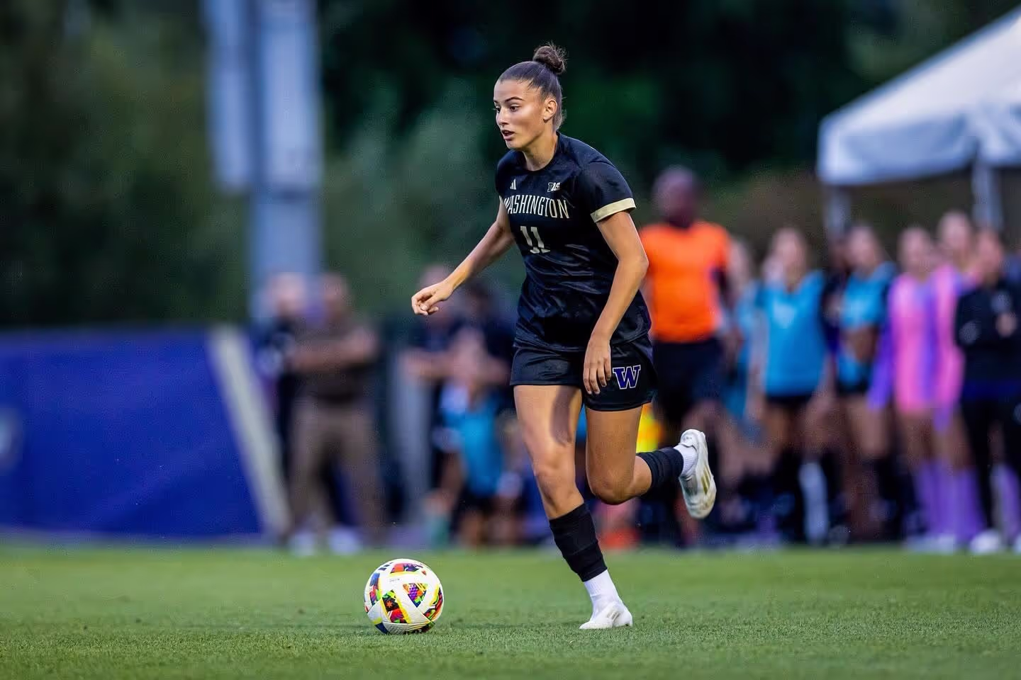 Female soccer player in black Washington uniform dribbling a colorful soccer ball on grass field.