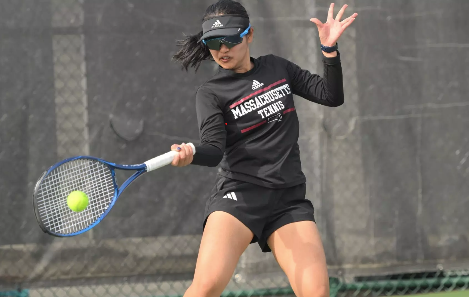 Female tennis player in black Massachusetts Tennis outfit hitting a forehand shot with a blue racquet and yellow ball in mid-air.