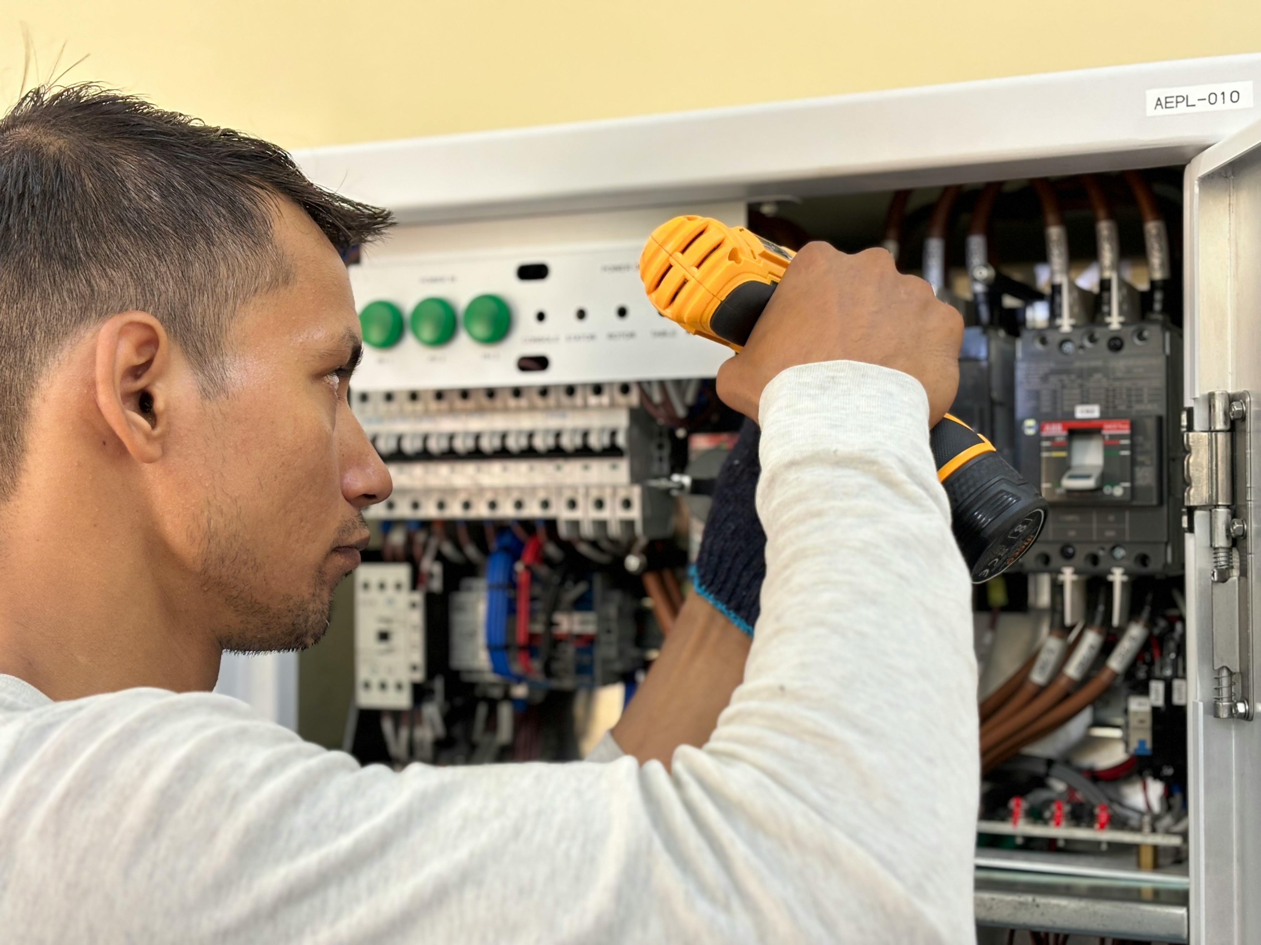 Electrician using a cordless drill to work on an electrical panel with multiple circuit breakers and wiring.
