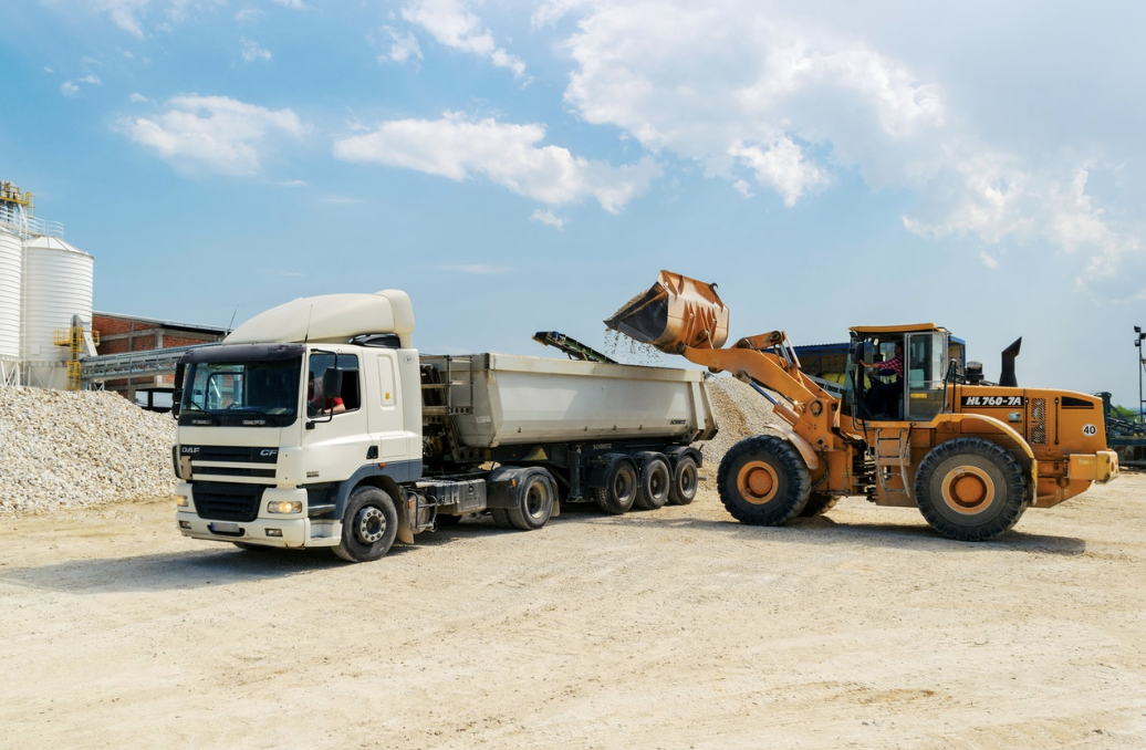 Orange excavator loading gravel into a white dump truck under a blue sky at a quarry site.