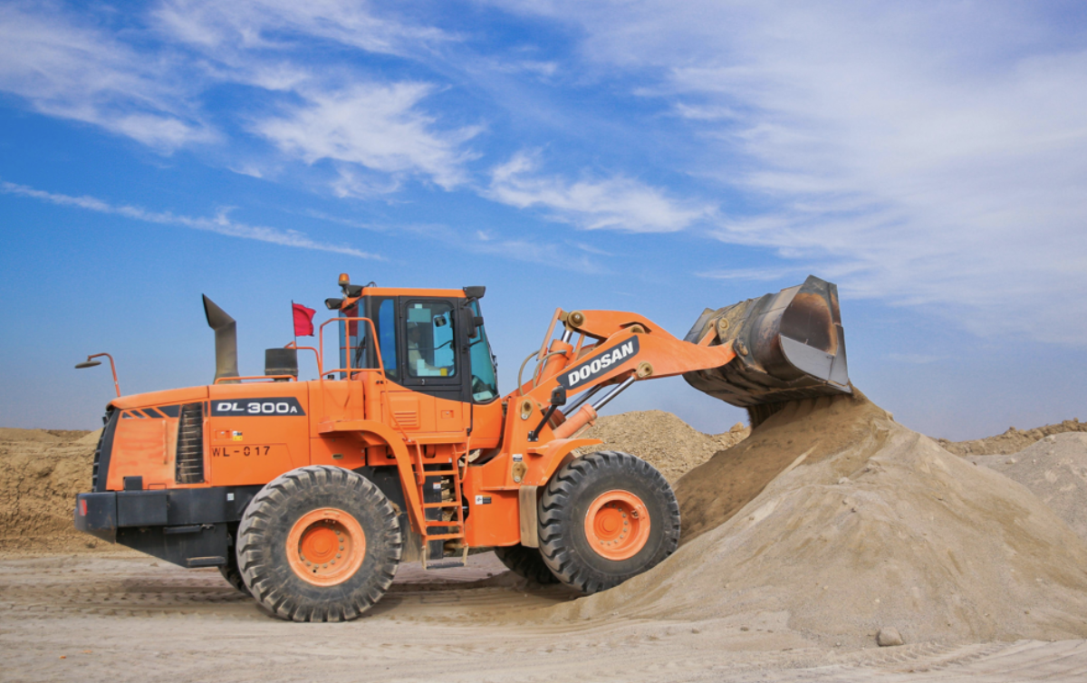 Orange Doosan DL300A loader moving a pile of sand at a construction site under a blue sky.