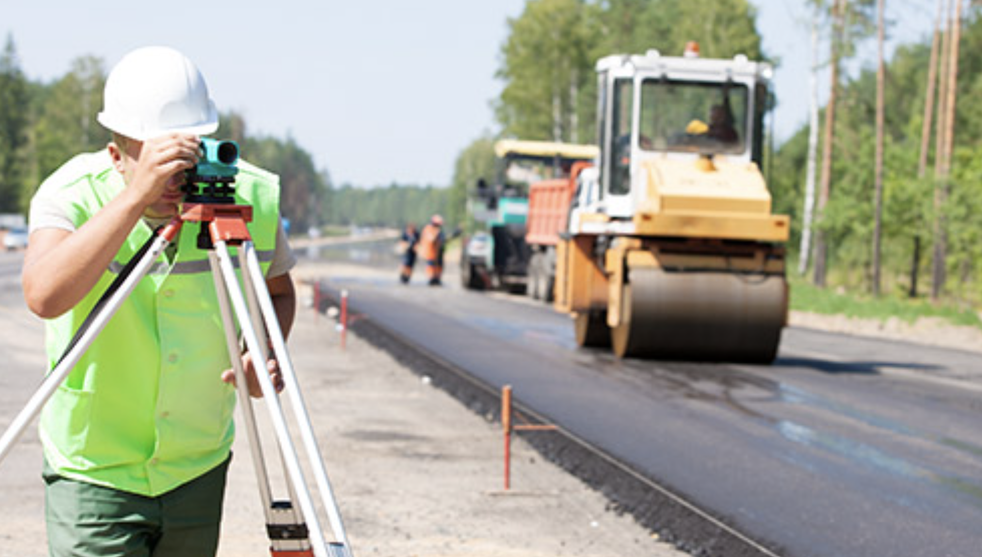 Construction worker in a white hard hat and neon green vest using a surveying instrument by a road under construction with heavy machinery.