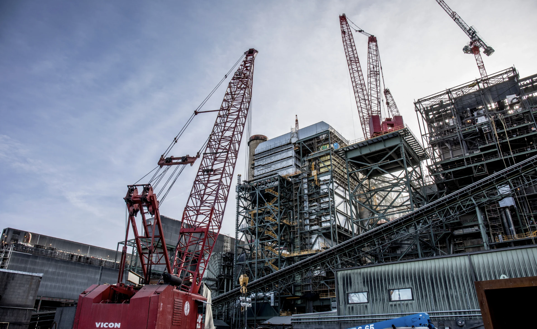 Large industrial construction site with red cranes and complex steel framework under a cloudy sky.