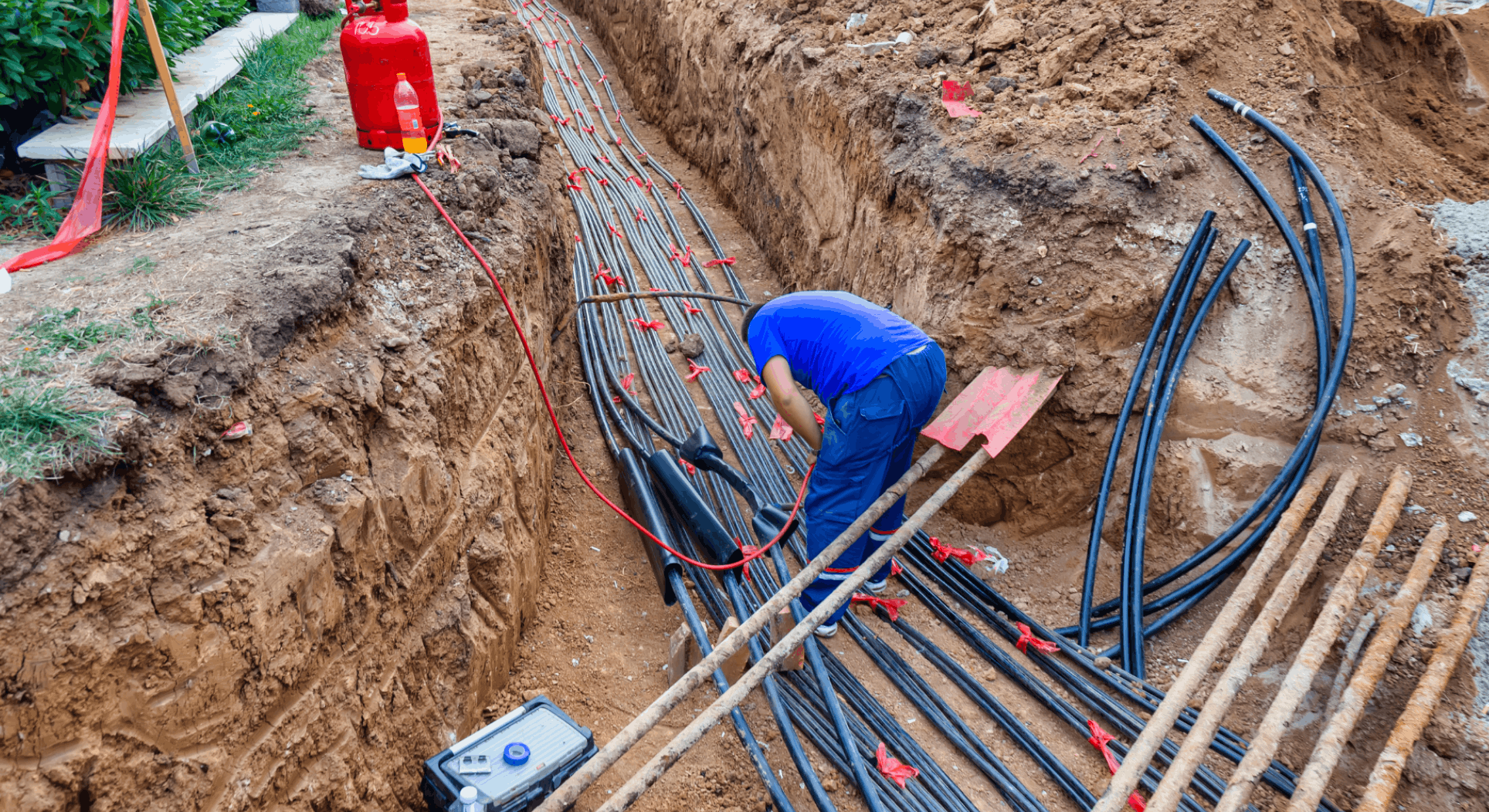 Worker in blue shirt installing underground electrical cables in a deep trench at a construction site.