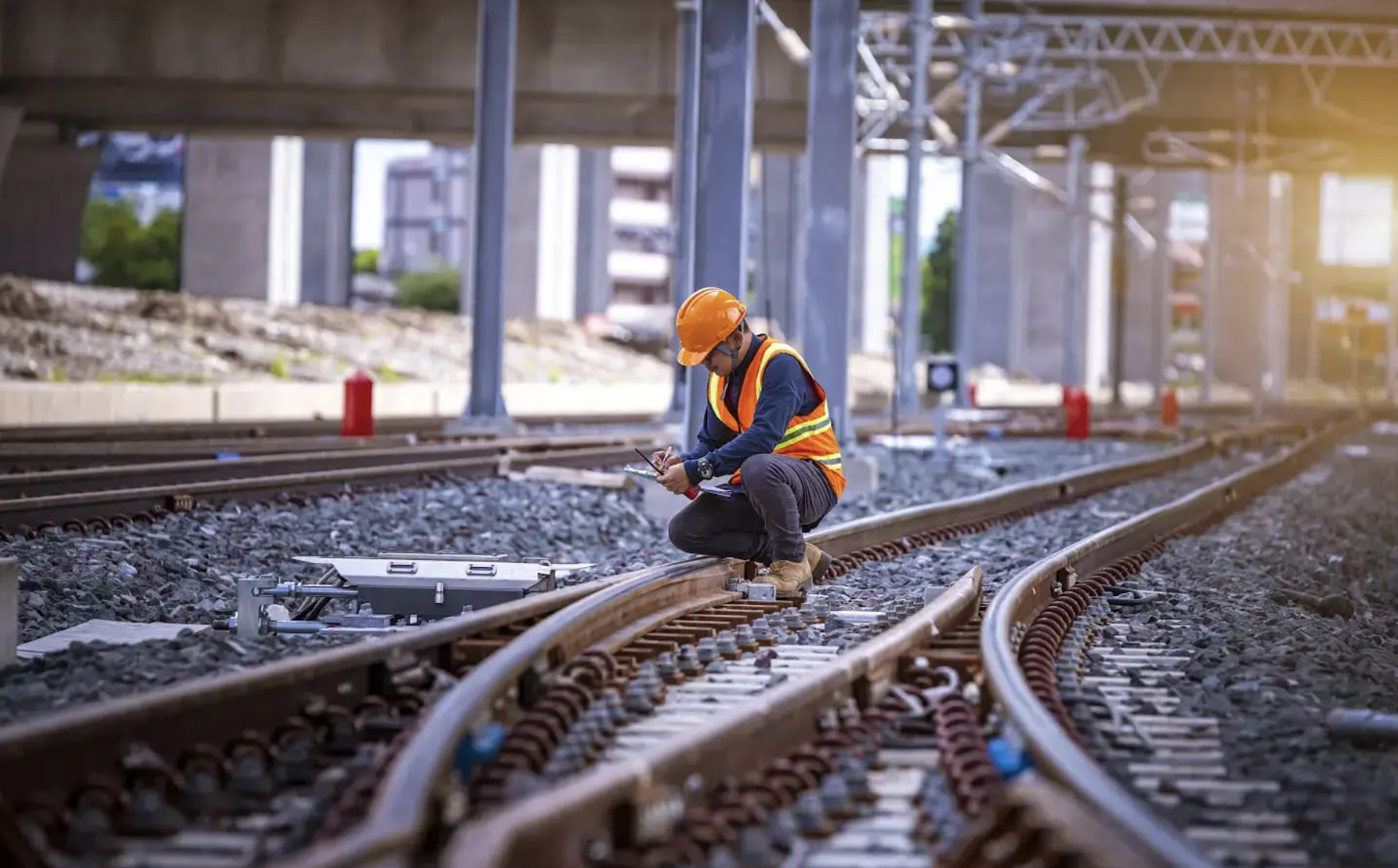 Railway worker in orange safety vest and helmet inspecting and taking notes on train tracks.