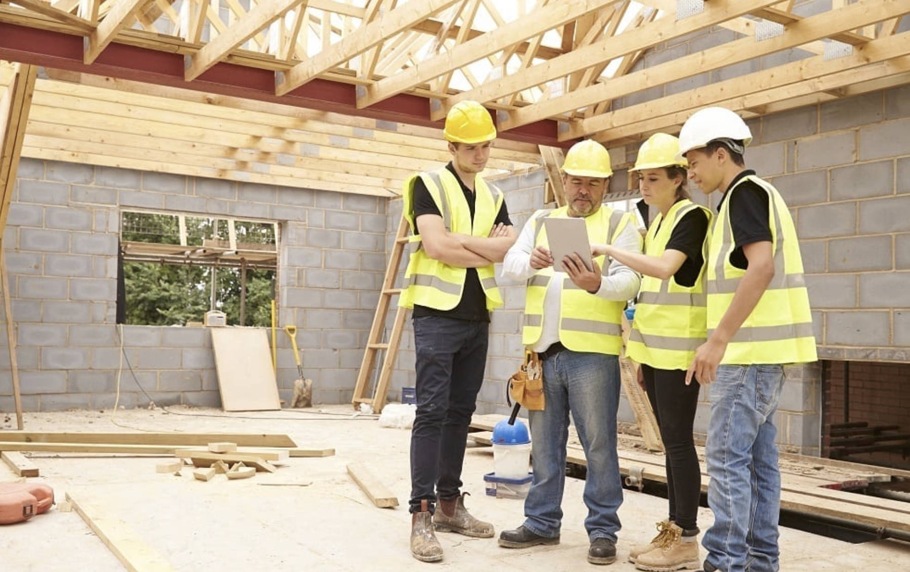 Four construction workers wearing safety vests and helmets reviewing a tablet inside a building under construction.