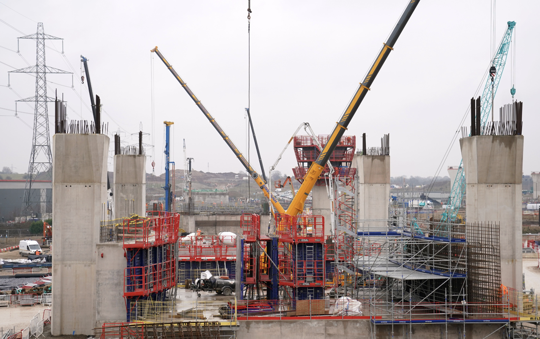 Construction site with large concrete pillars, cranes, scaffolding, and workers building a bridge or elevated structure.