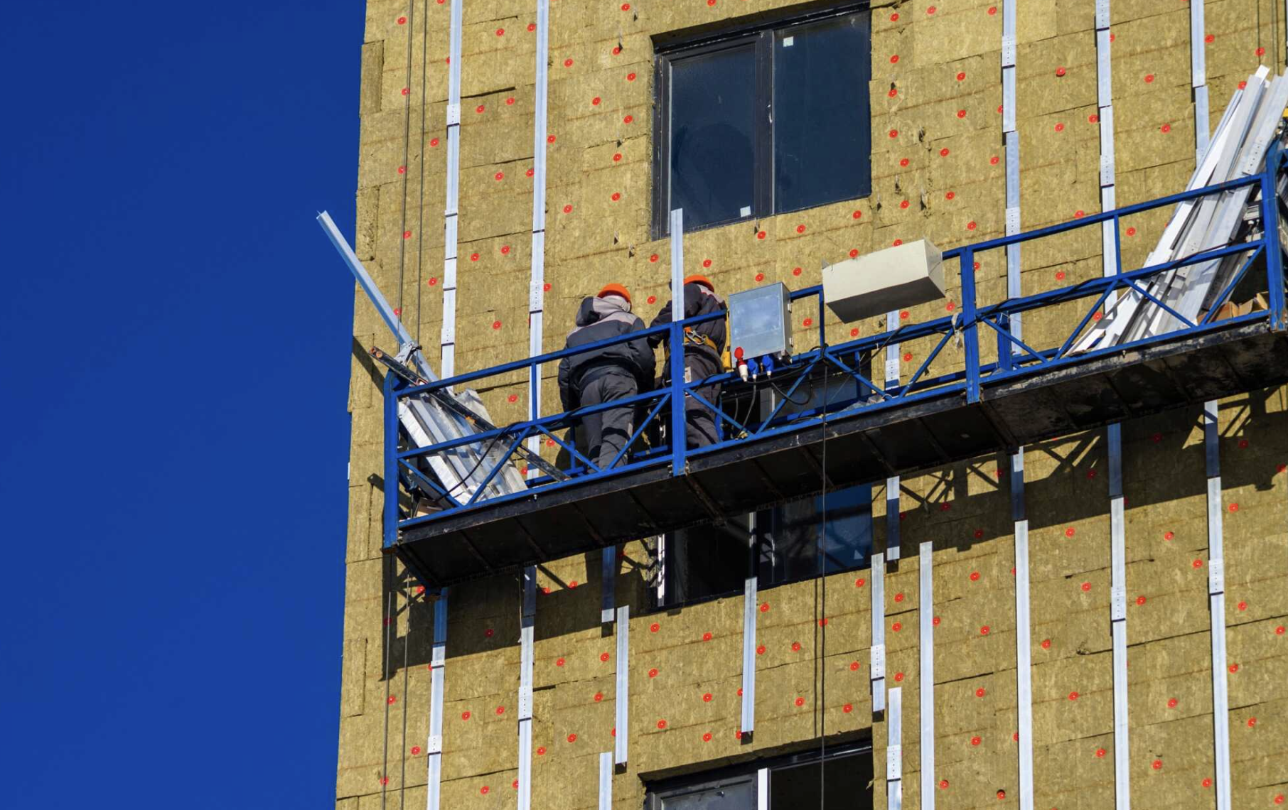 Two construction workers in safety helmets working on a suspended scaffold on a building facade with insulation panels and metal strips.