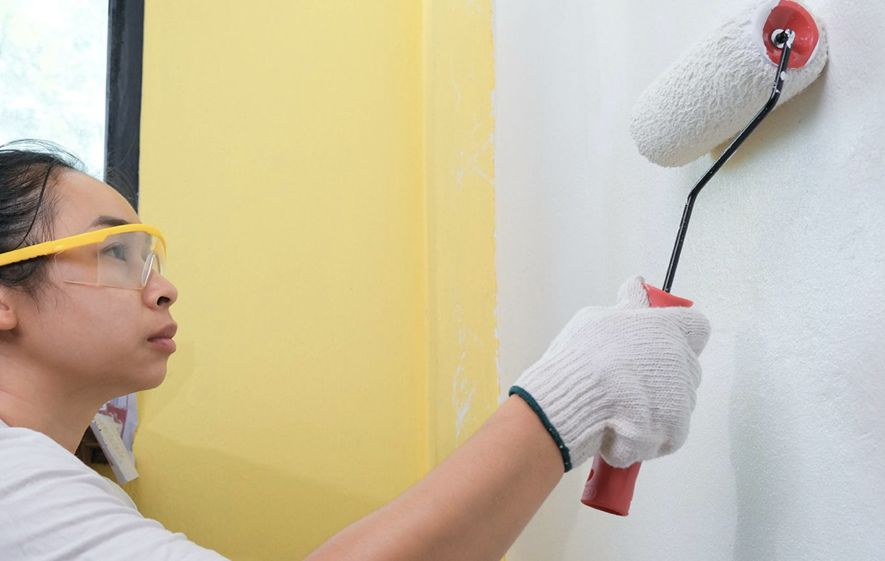 Person wearing safety glasses and gloves painting a wall white with a paint roller, next to a yellow painted wall.