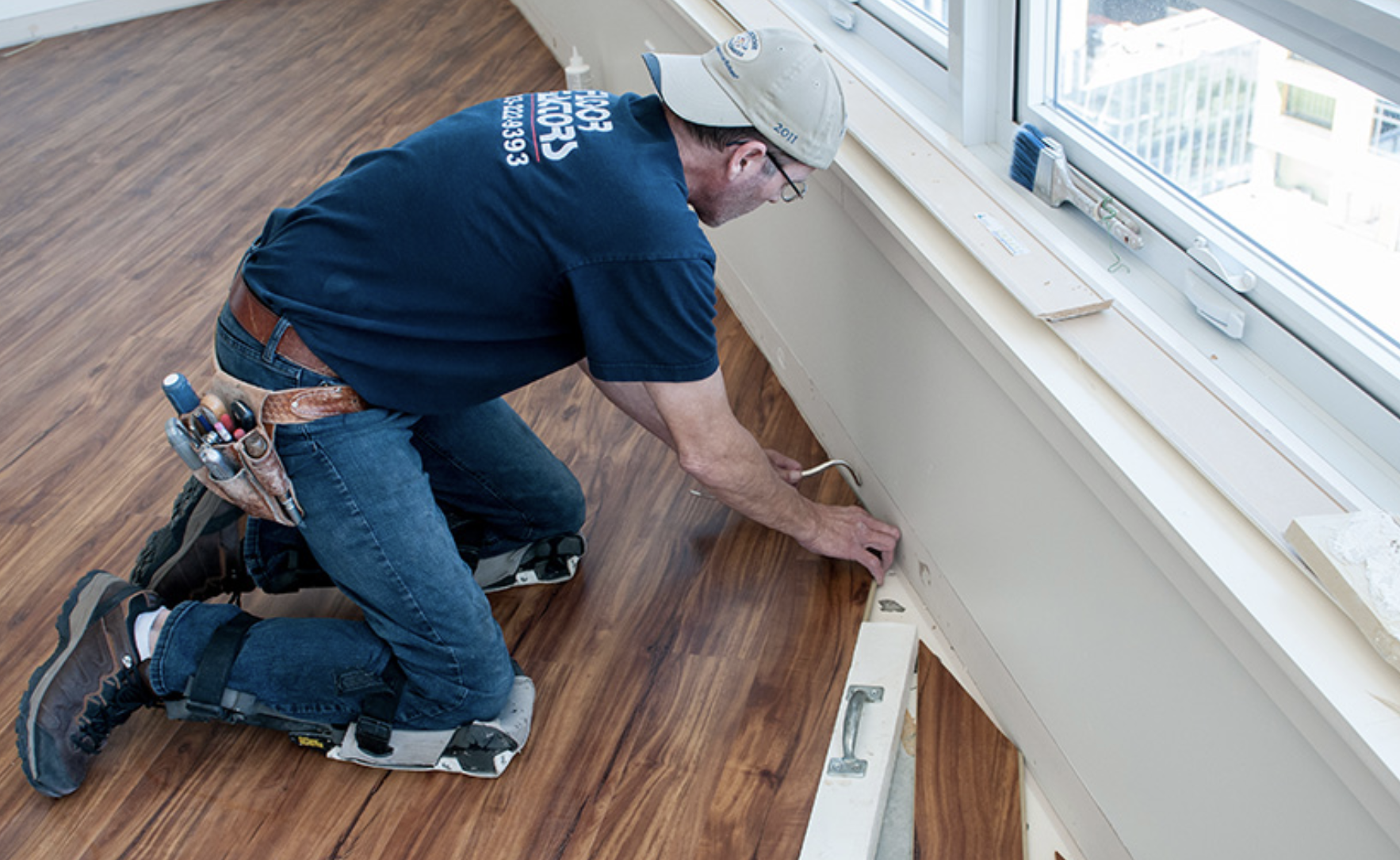 A man wearing knee pads and a tool belt installing wooden flooring near a window.