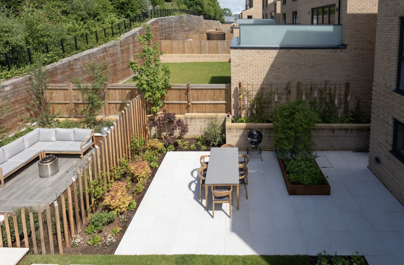 Modern backyard patio with outdoor dining table and chairs, a grill, garden beds, and a cushioned outdoor sofa on a wooden deck.