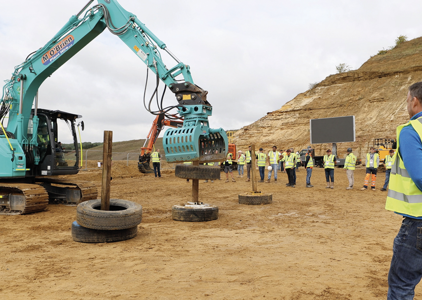 Construction site with a turquoise excavator lifting a tire around a wooden post while workers in high-visibility vests observe.