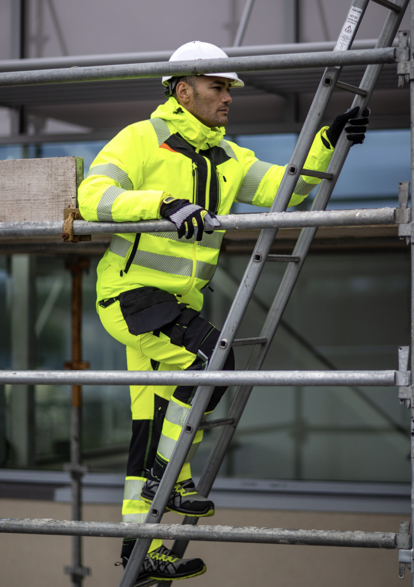 Construction worker in high-visibility yellow safety clothing and white helmet climbing a metal ladder on scaffolding.