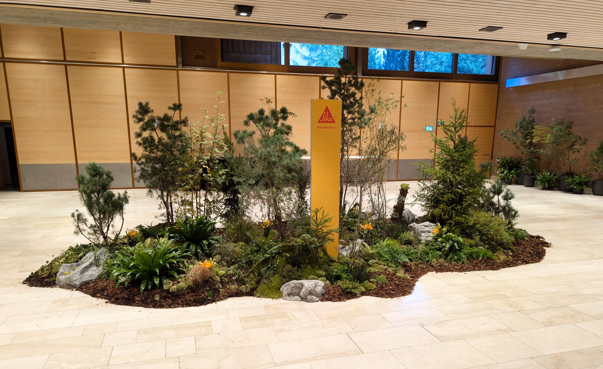 Indoor garden with small pine trees, green plants, rocks, and a yellow sign with the Sika logo in a large room with wooden panel walls and tiled floor.