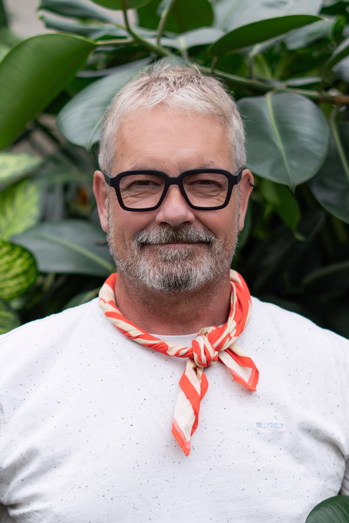 Smiling middle-aged man with gray hair, black glasses, and a red and white scarf, standing in front of large green leaves.