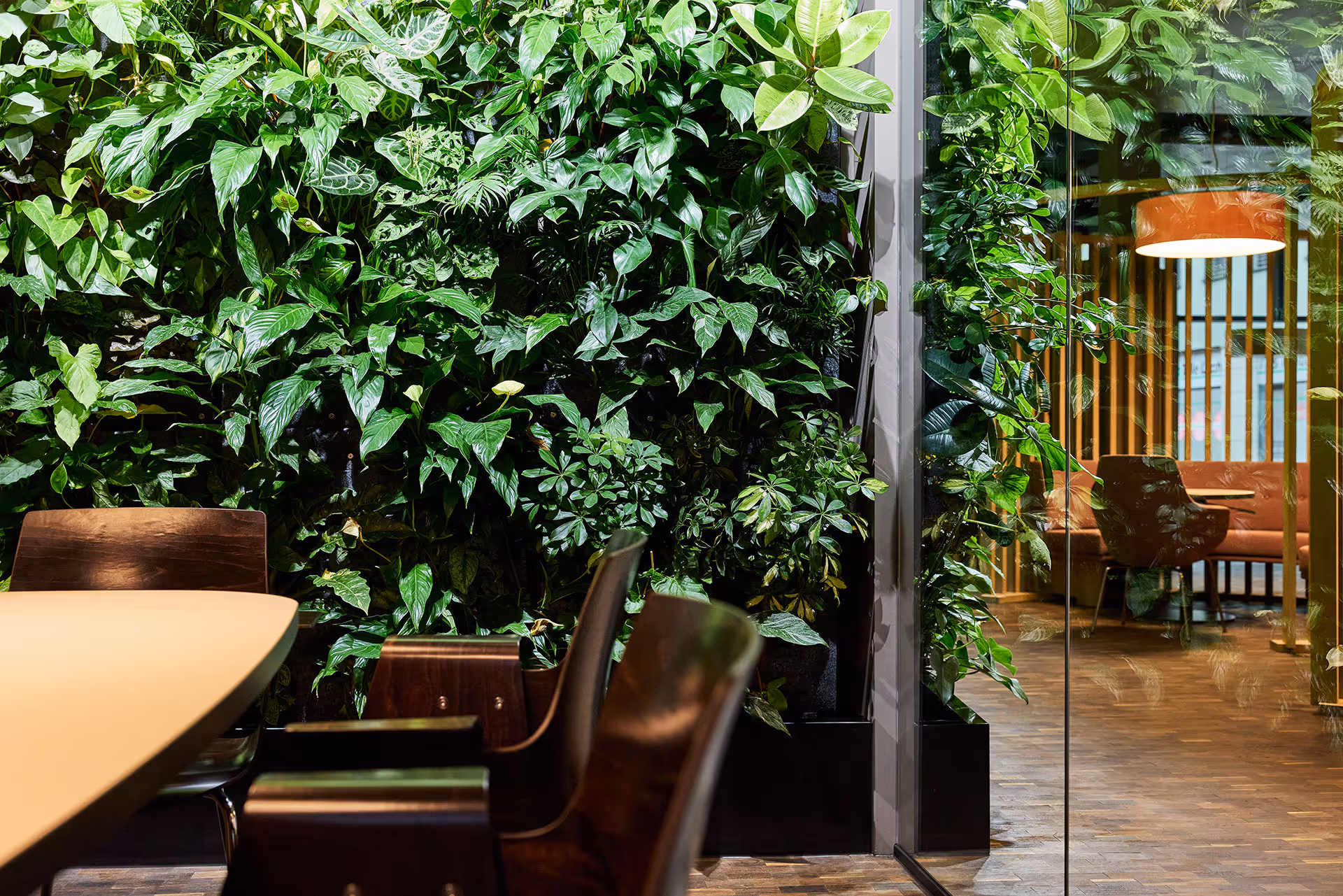 Indoor office space with a green vertical plant wall, wooden chairs around a table, and a glass partition revealing a lounge area with an orange pendant light.