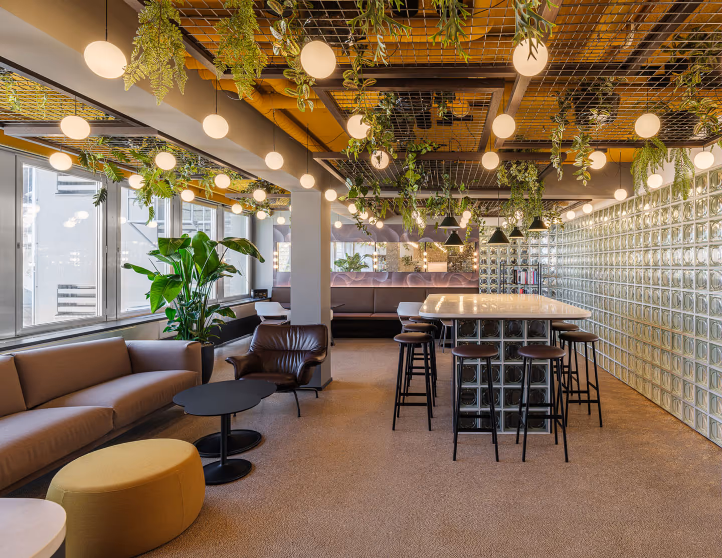 Modern office lounge with a beige sofa, brown leather armchair, black coffee tables, green plants, hanging round pendant lights, and a high table with stools.