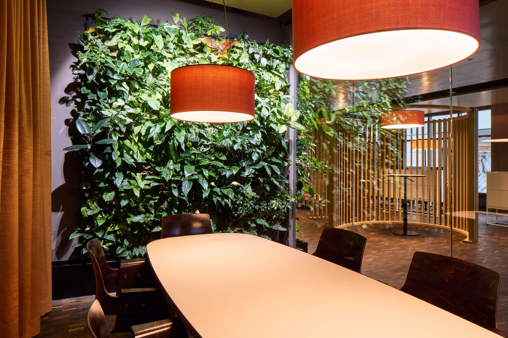 Modern meeting room with a long white table, dark chairs, hanging orange lamps, and a green plant wall.