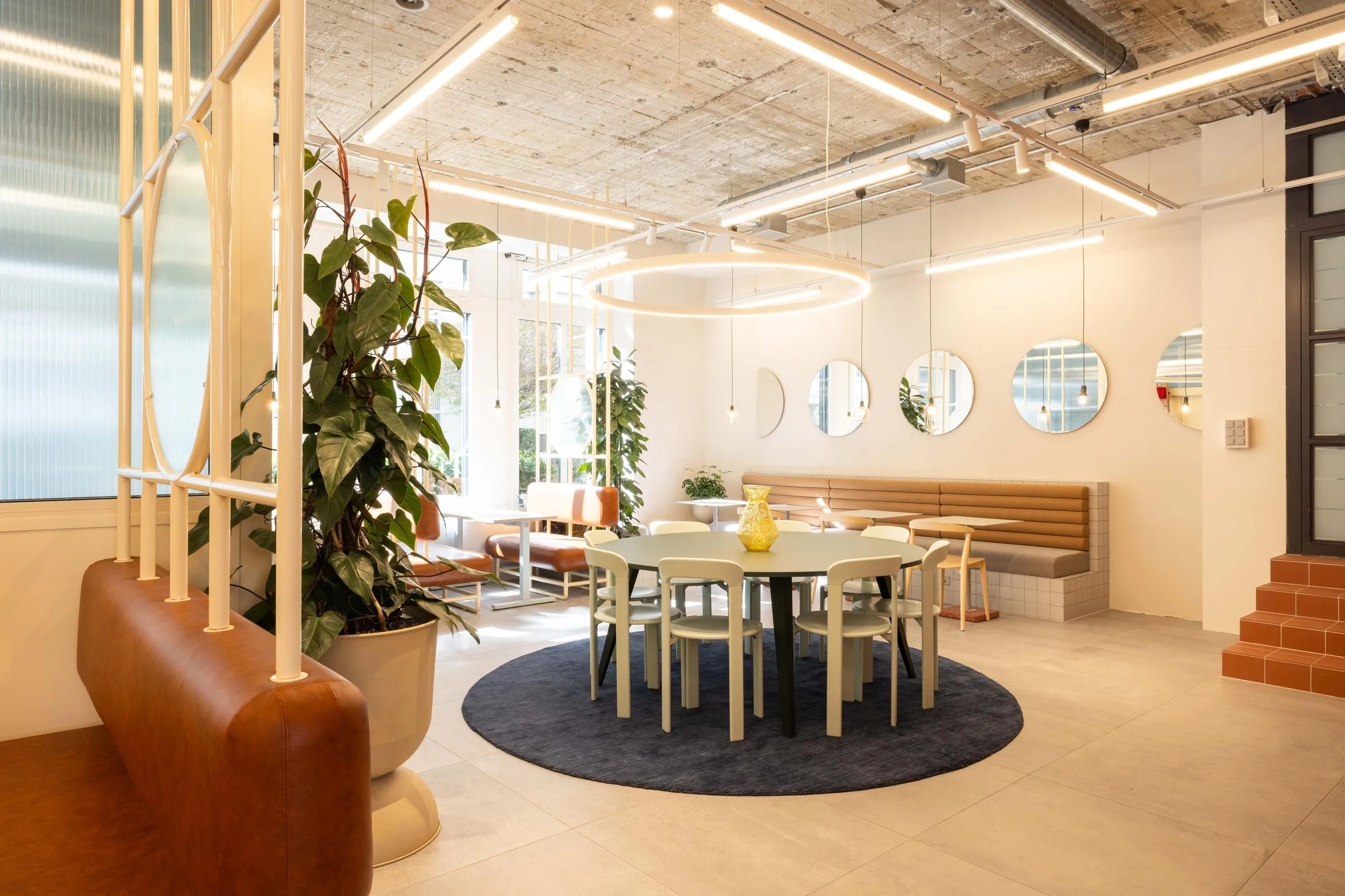 Bright modern office lounge with circular table, white chairs, blue rug, brown leather bench, plants, and round mirrors on the wall.