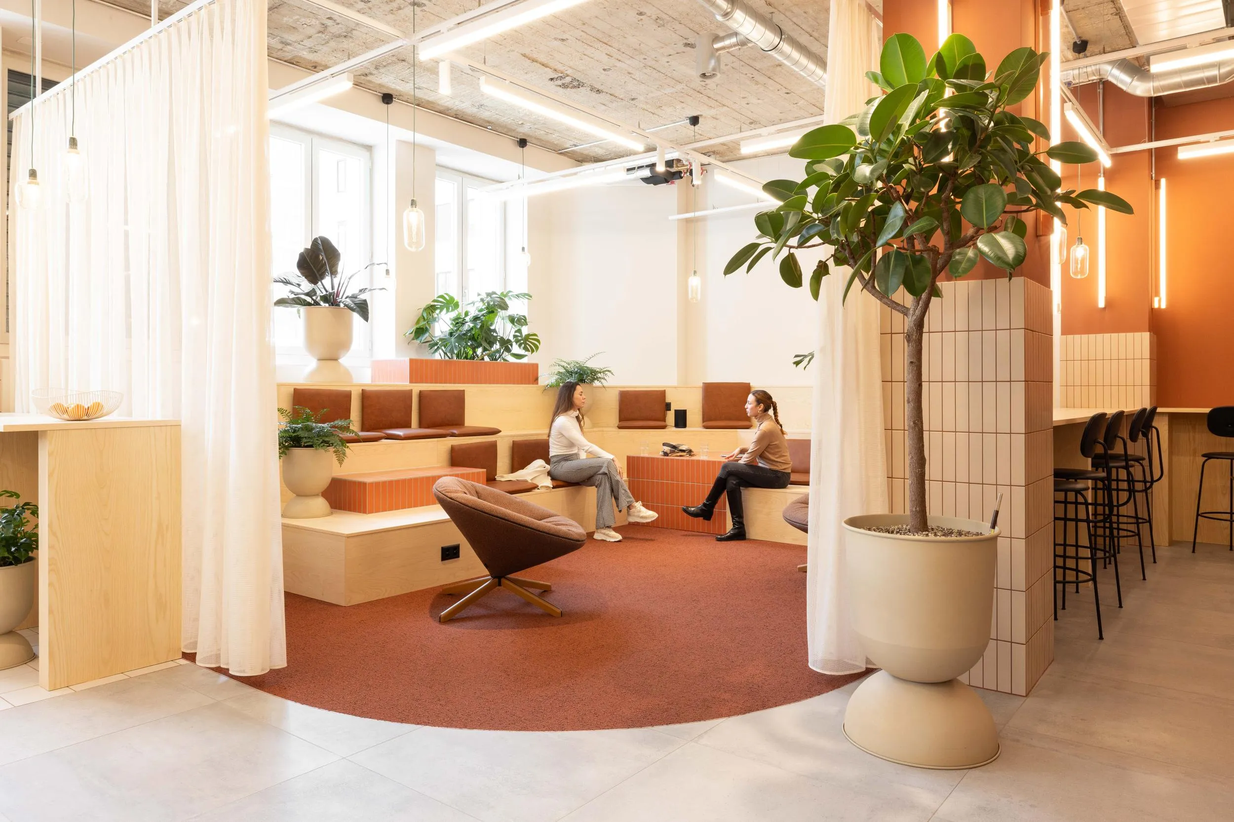 Modern office lounge with tiered seating, two women talking, large plants, and warm lighting.