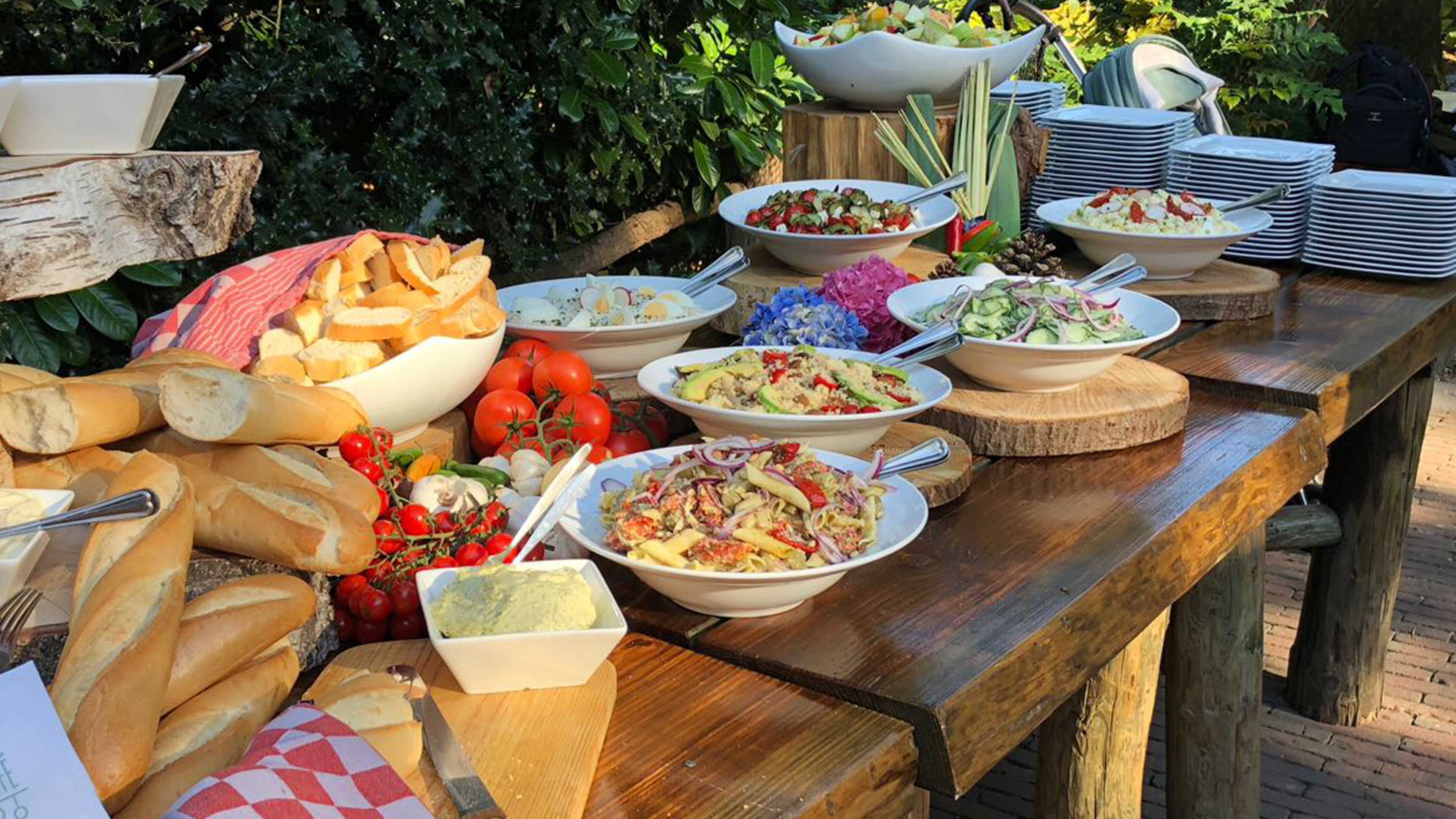 Buitenbuffet met salades, stokbrood en tomaten op houten tafels in de tuin van De Baron