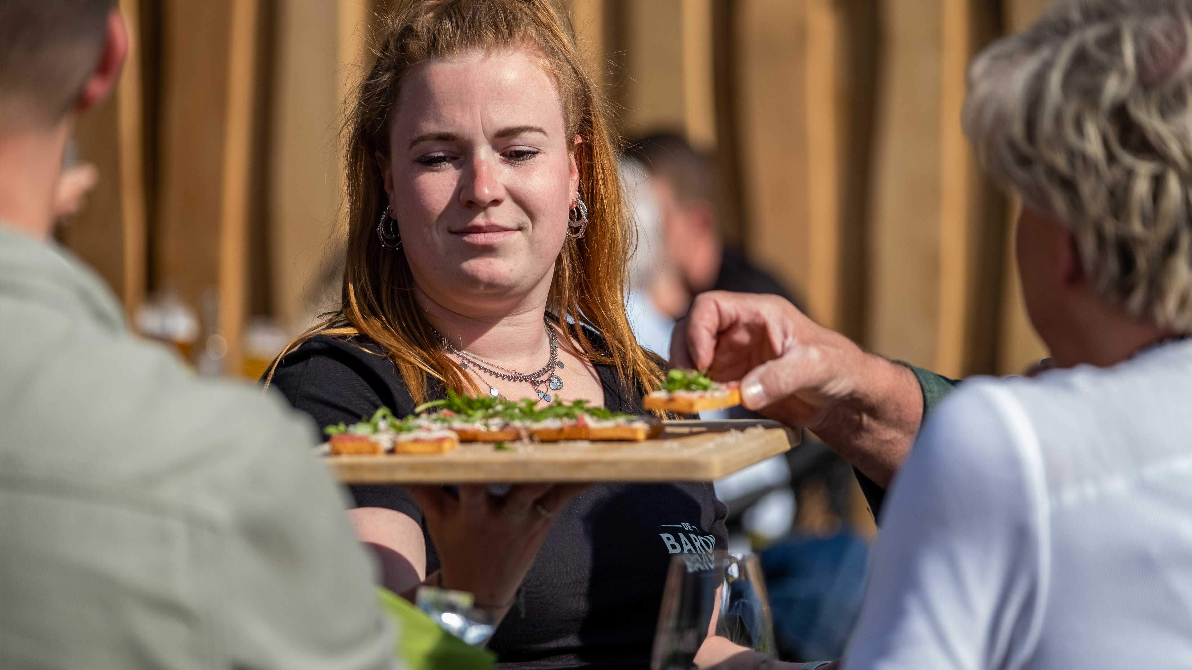 Medewerker van De Baron serveert hapjes op een plank aan gasten op het terras