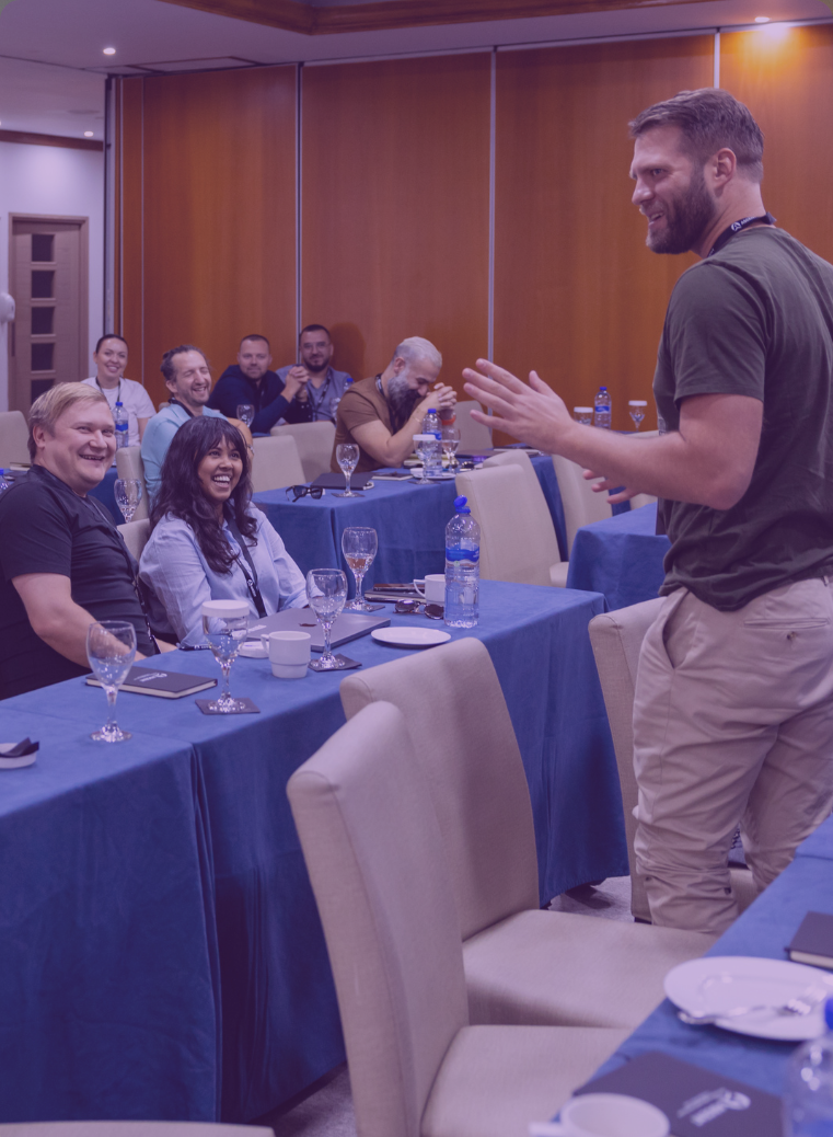 Group of people mingling and conversing in a bright indoor space with white walls and a wooden table with chairs in the foreground.