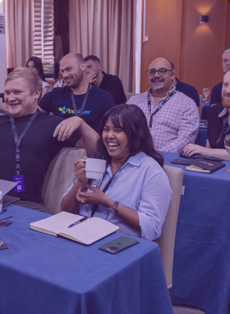 Group of diverse adults smiling and laughing in a meeting room with notebooks and drinks on tables covered by blue cloths.