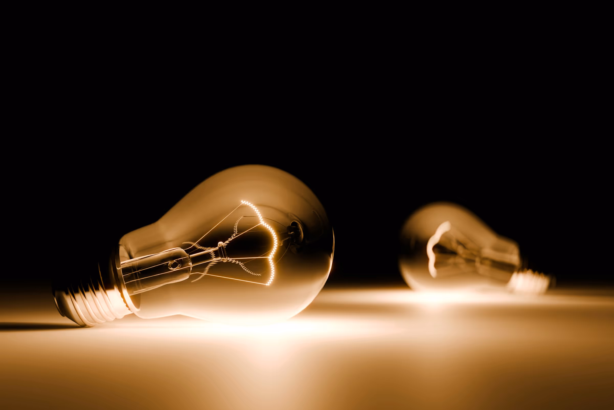 Two incandescent light bulbs lying on a surface, one in sharp focus and glowing warmly, the other blurred in the background.