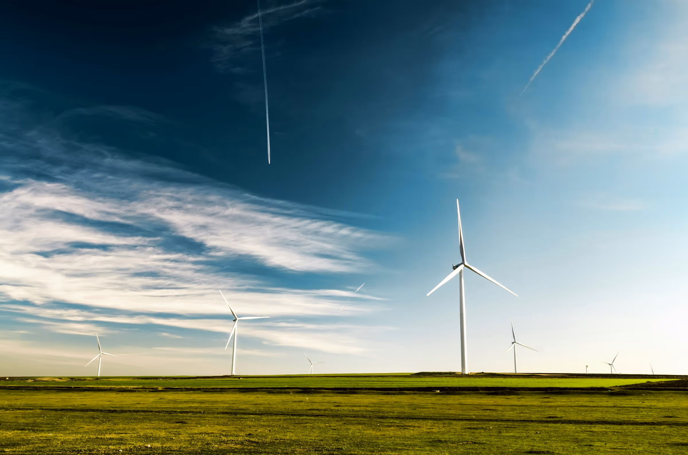 Wind turbines on a green field under a blue sky with scattered clouds and airplane contrails.