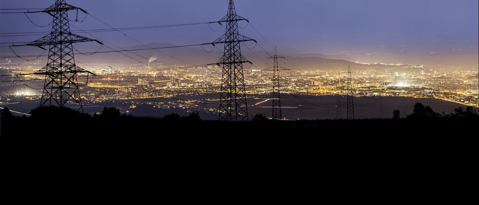 Cityscape at dusk with illuminated buildings and multiple tall electrical transmission towers in the foreground.