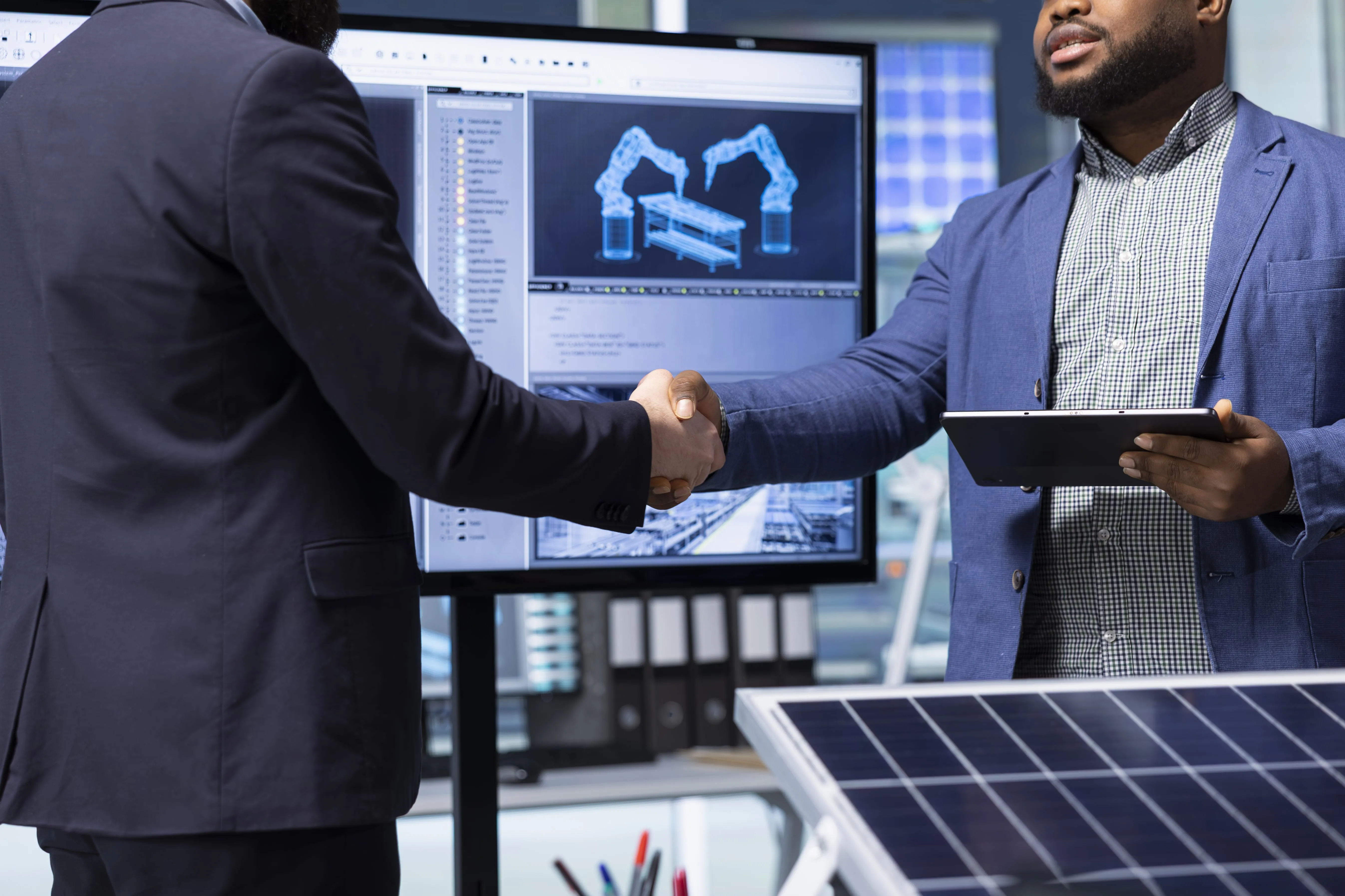 Two men in business attire shaking hands in front of a screen showing robotic arm designs and a solar panel in the foreground.