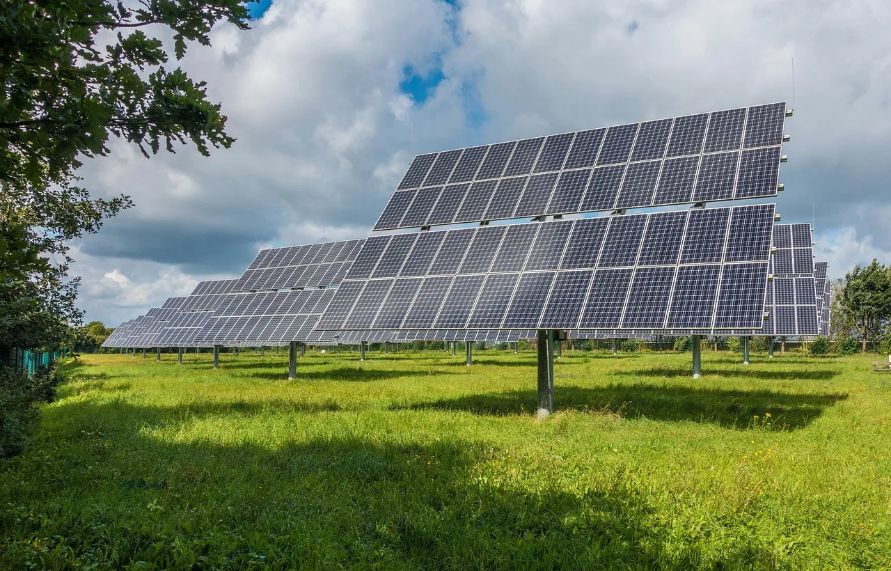 Multiple solar panels installed on metal stands in a green grassy field under a partly cloudy sky.