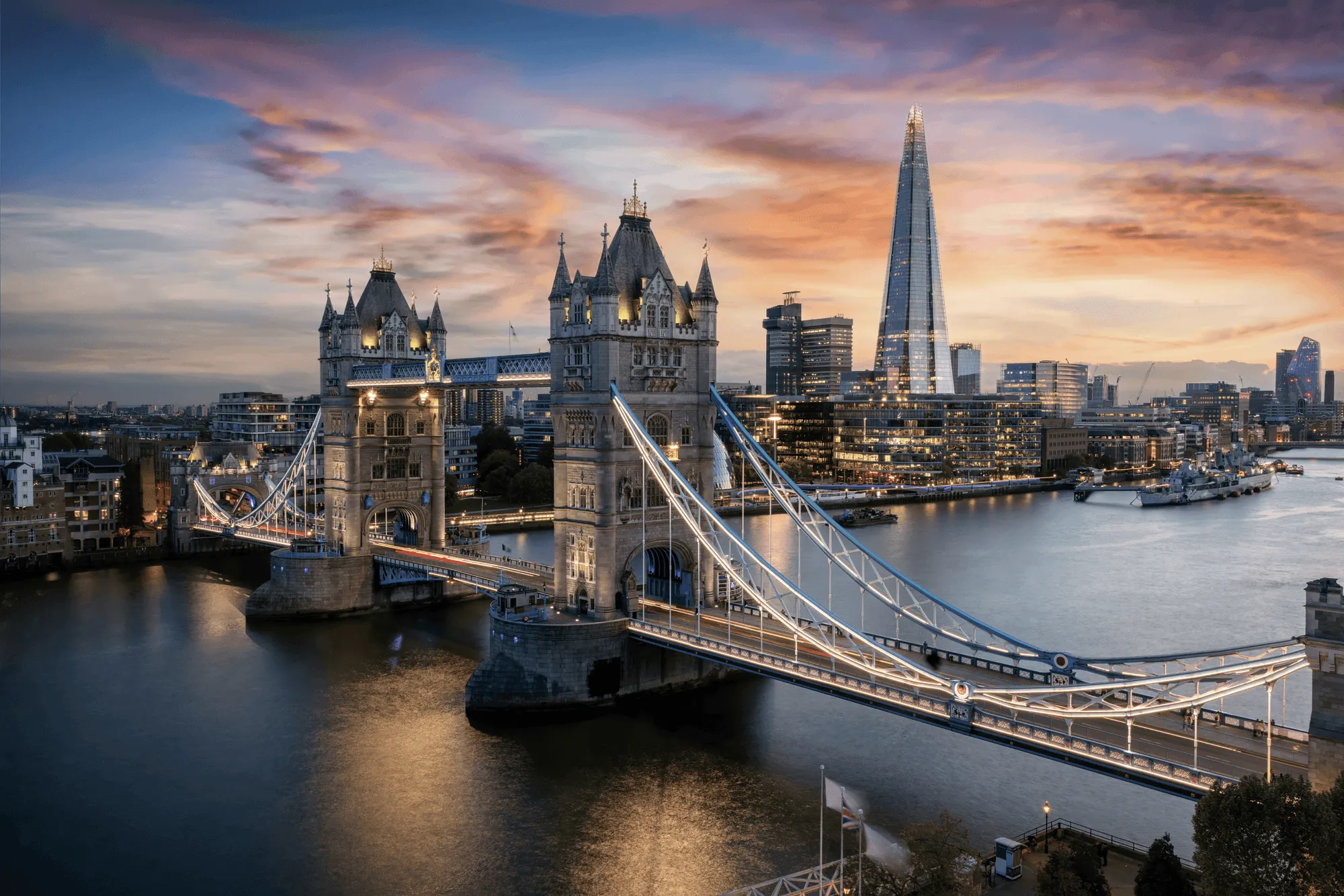 Illuminated Tower Bridge over the River Thames at dusk with the Shard skyscraper and city buildings in the background.