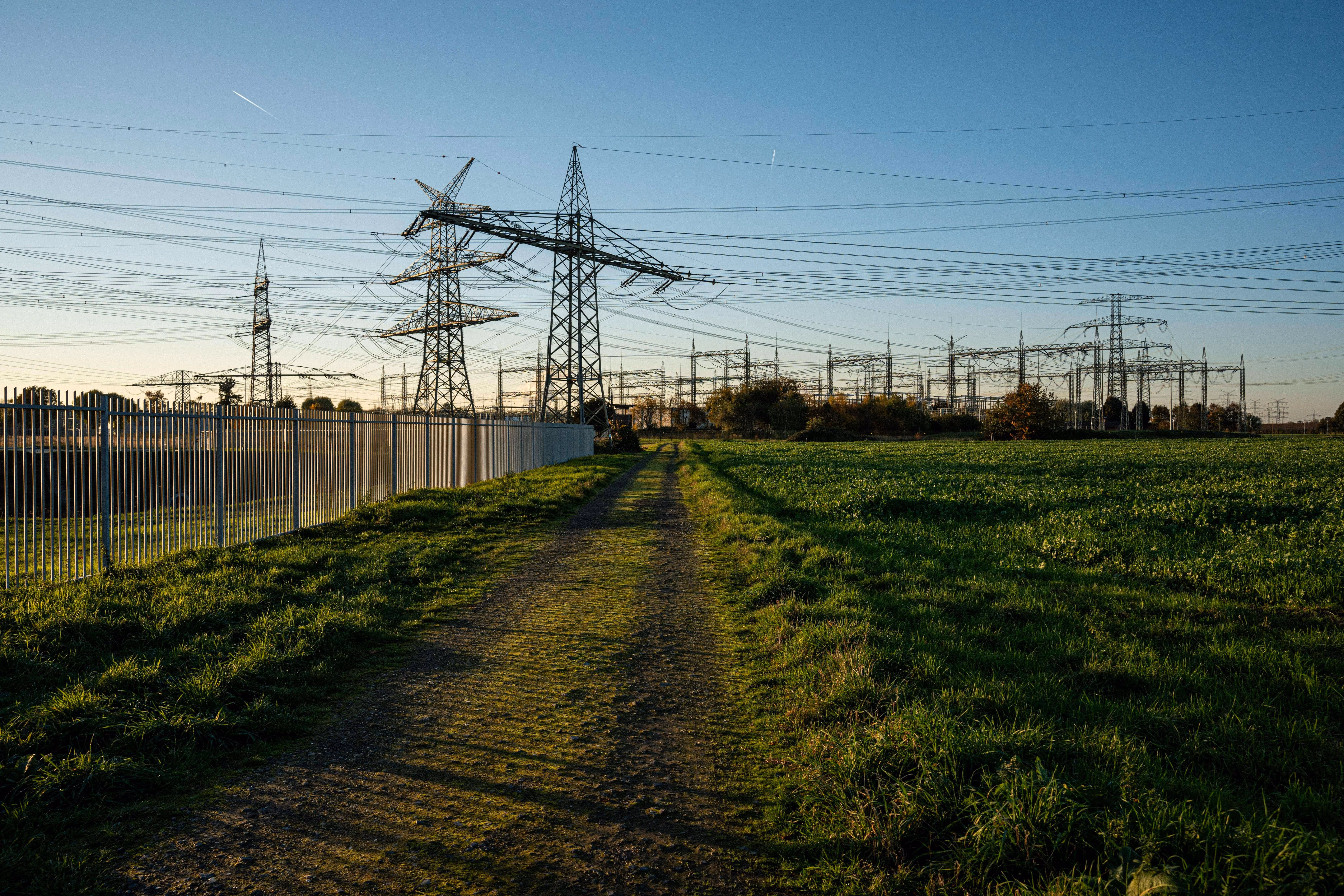 Dirt path between green fields leading to electrical power transmission towers and lines under a clear blue sky.